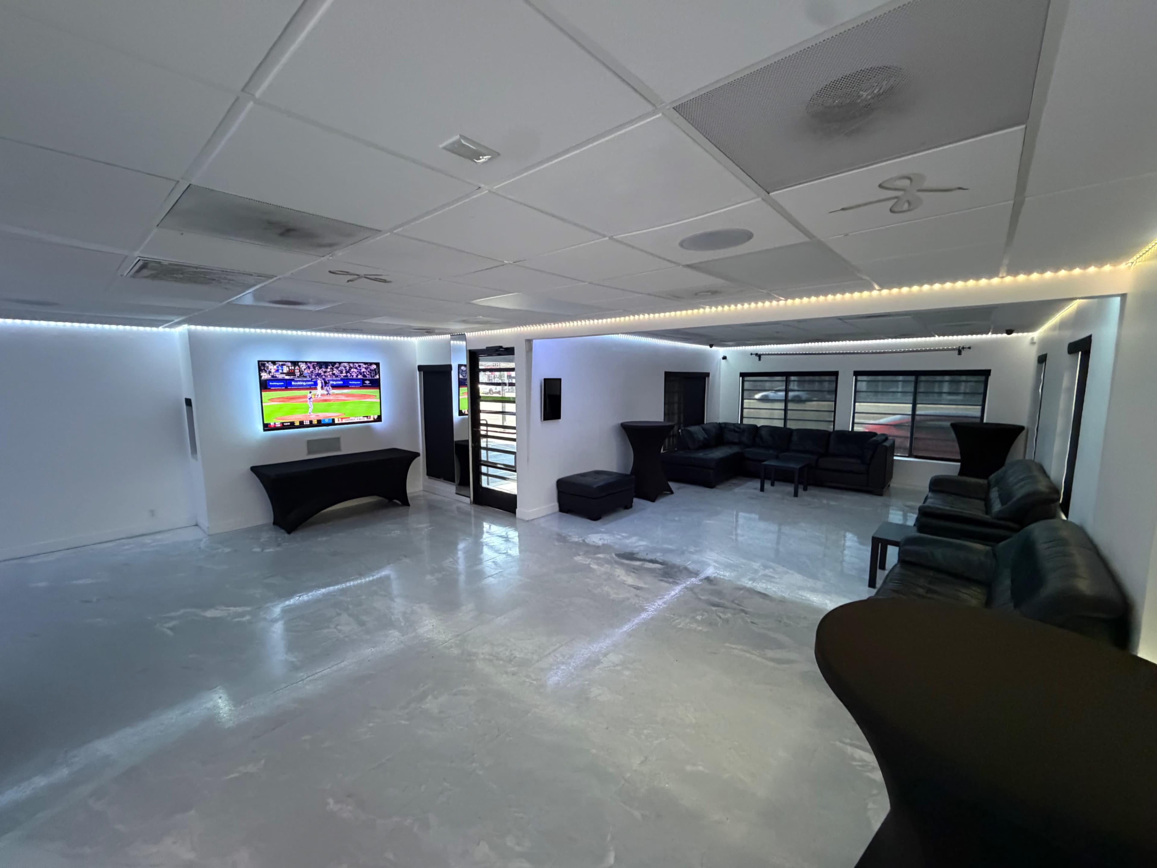 The image shows a modern lounge area with a large television displaying a sports game, black leather seating, and sleek black tables.