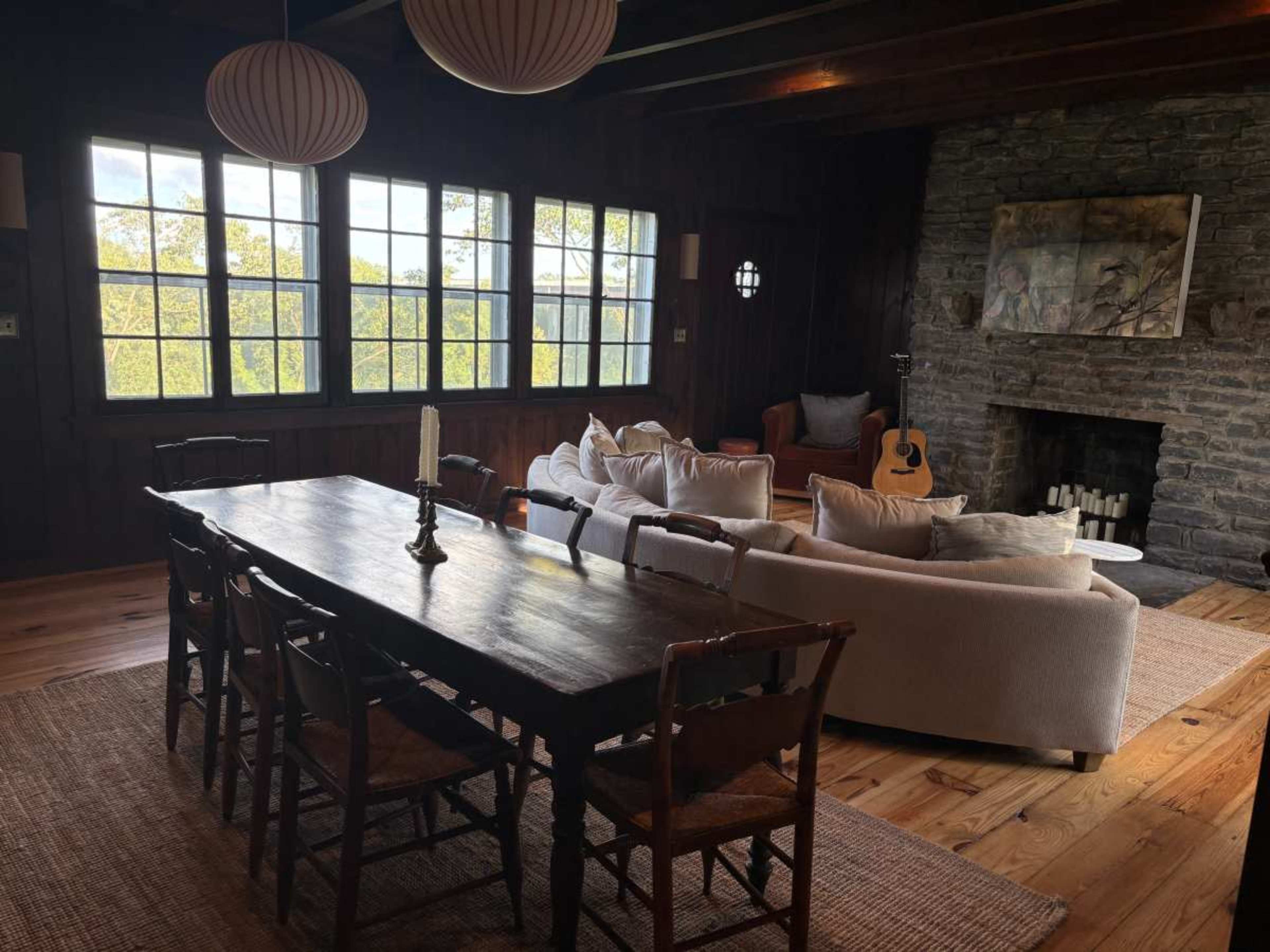 The image shows a rustic living space featuring a wooden dining table, a cozy couch, a stone fireplace, large windows with views of greenery, and an acoustic guitar resting nearby.