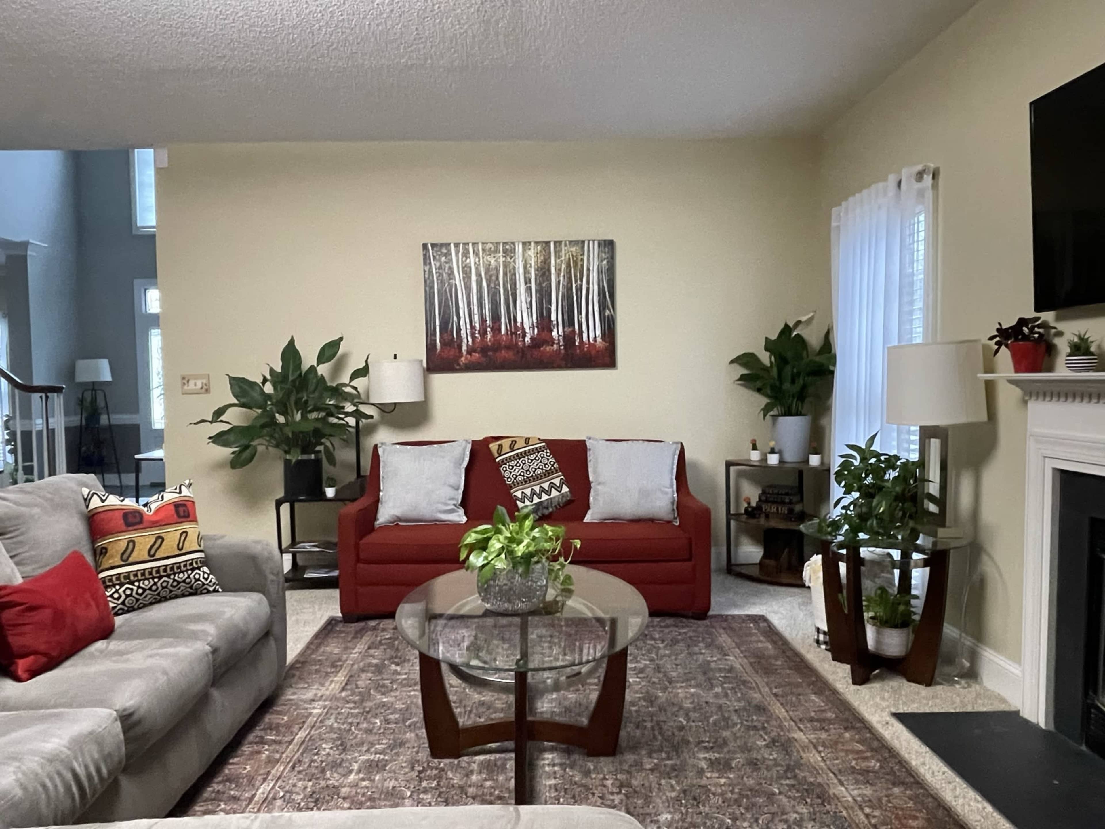 A living room with a red sofa, a glass coffee table, two potted plants, and a painting of trees on the wall.