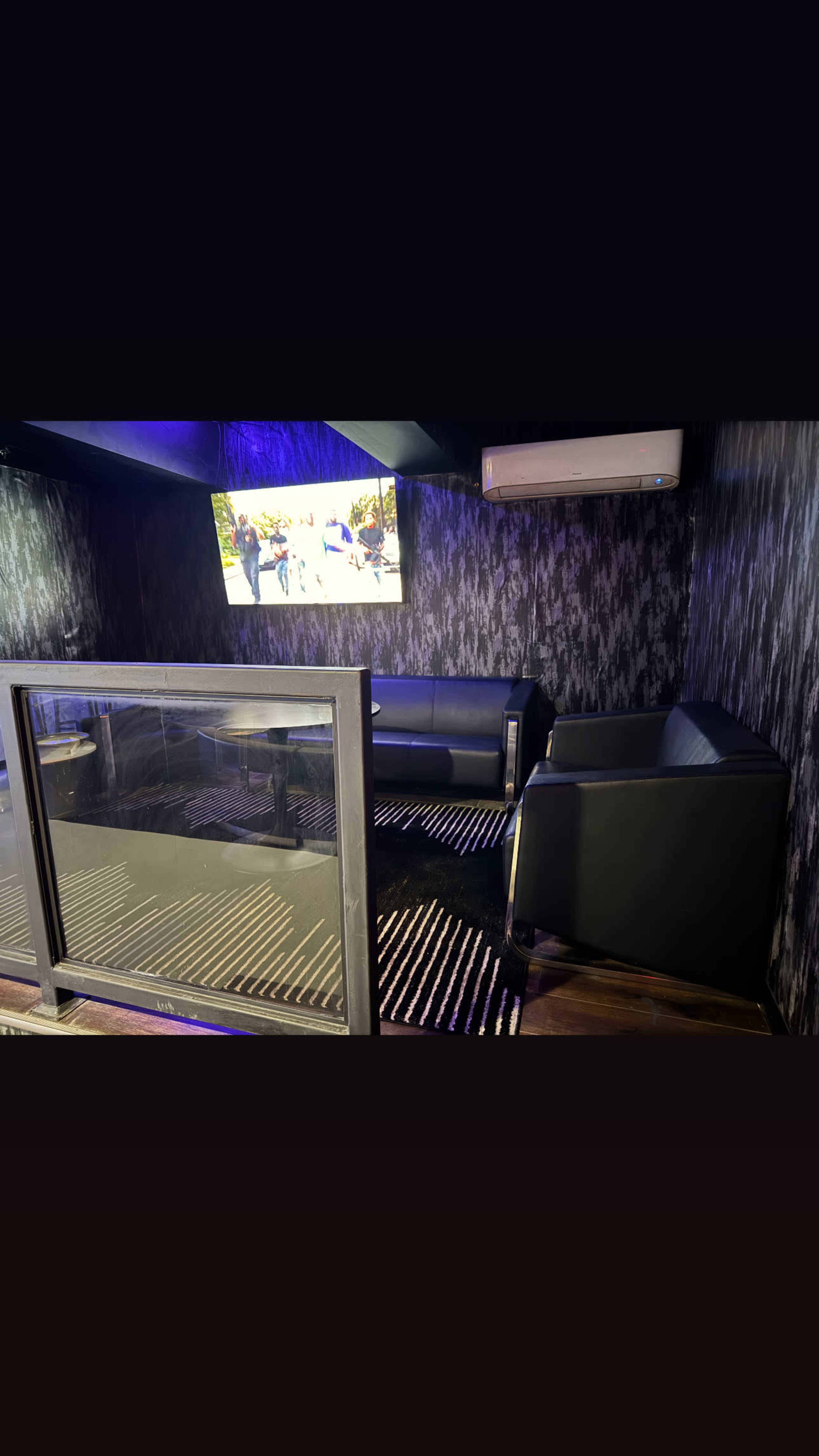 The image shows a contemporary lounge area with black seating, a table, and a wall-mounted screen in a dimly lit space.