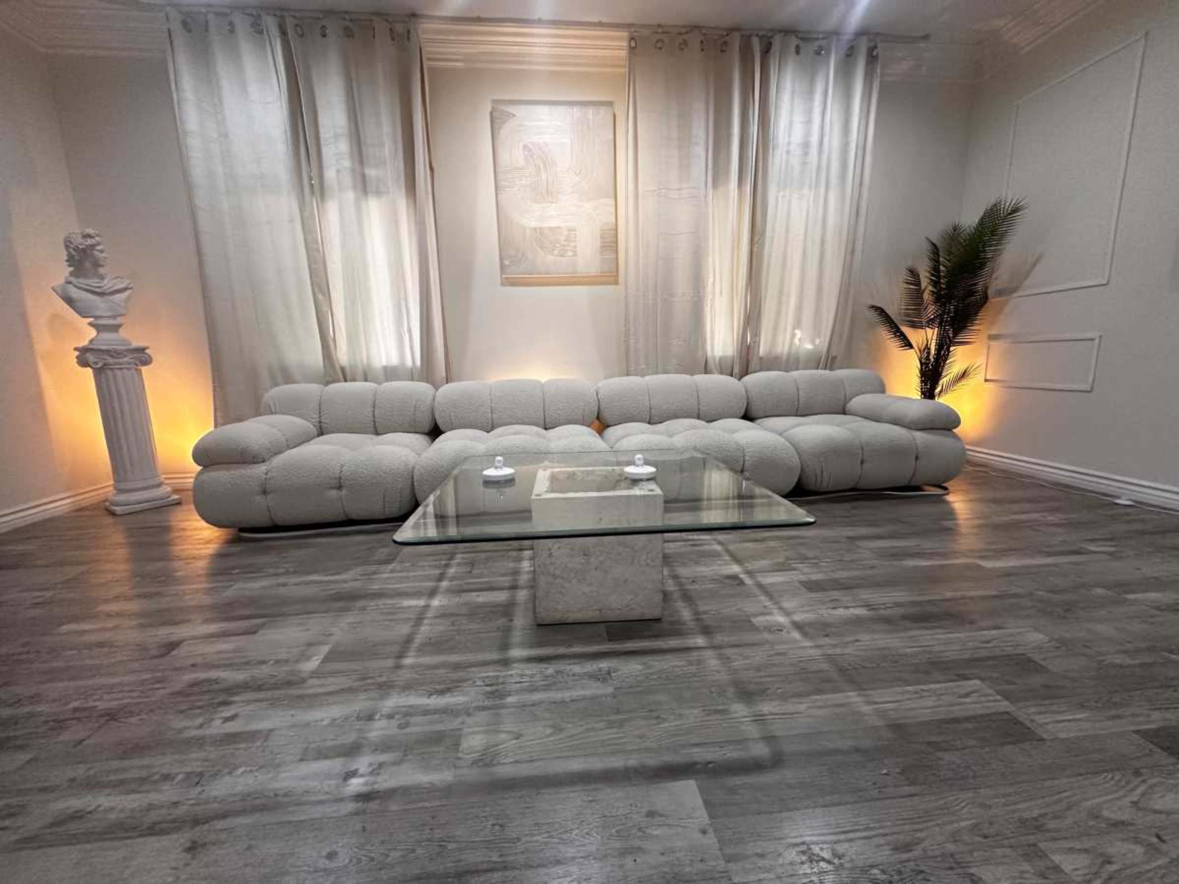 The image shows a modern, minimalist living room featuring a white, curved sofa, a glass coffee table, and a decorative statue with soft lighting from the walls.
