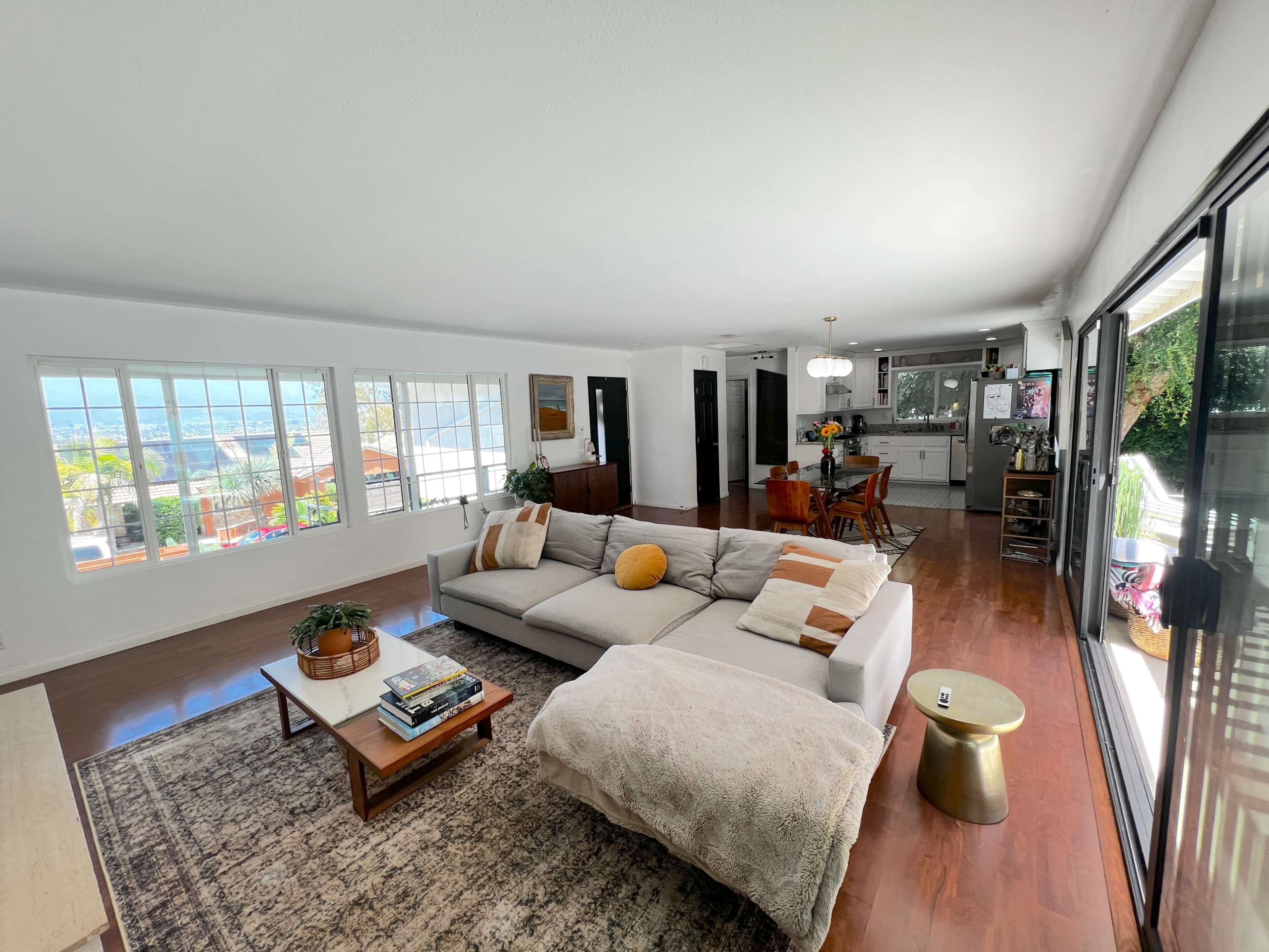 The living room features a large sectional sofa, a coffee table, and large windows that provide a view of the outdoors, with a dining area visible in the background.