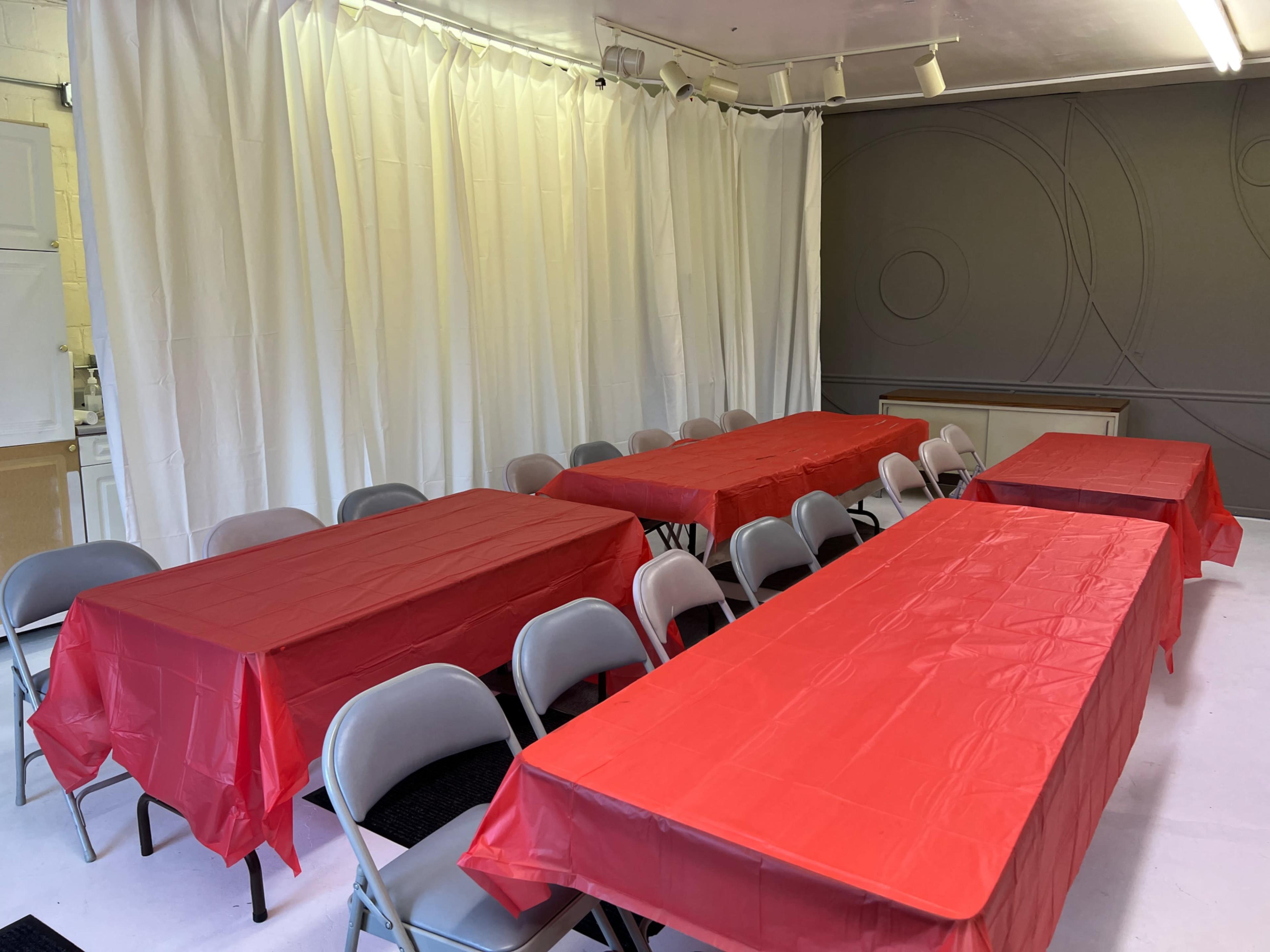 A room set up with several long tables covered in red tablecloths and surrounded by gray folding chairs.