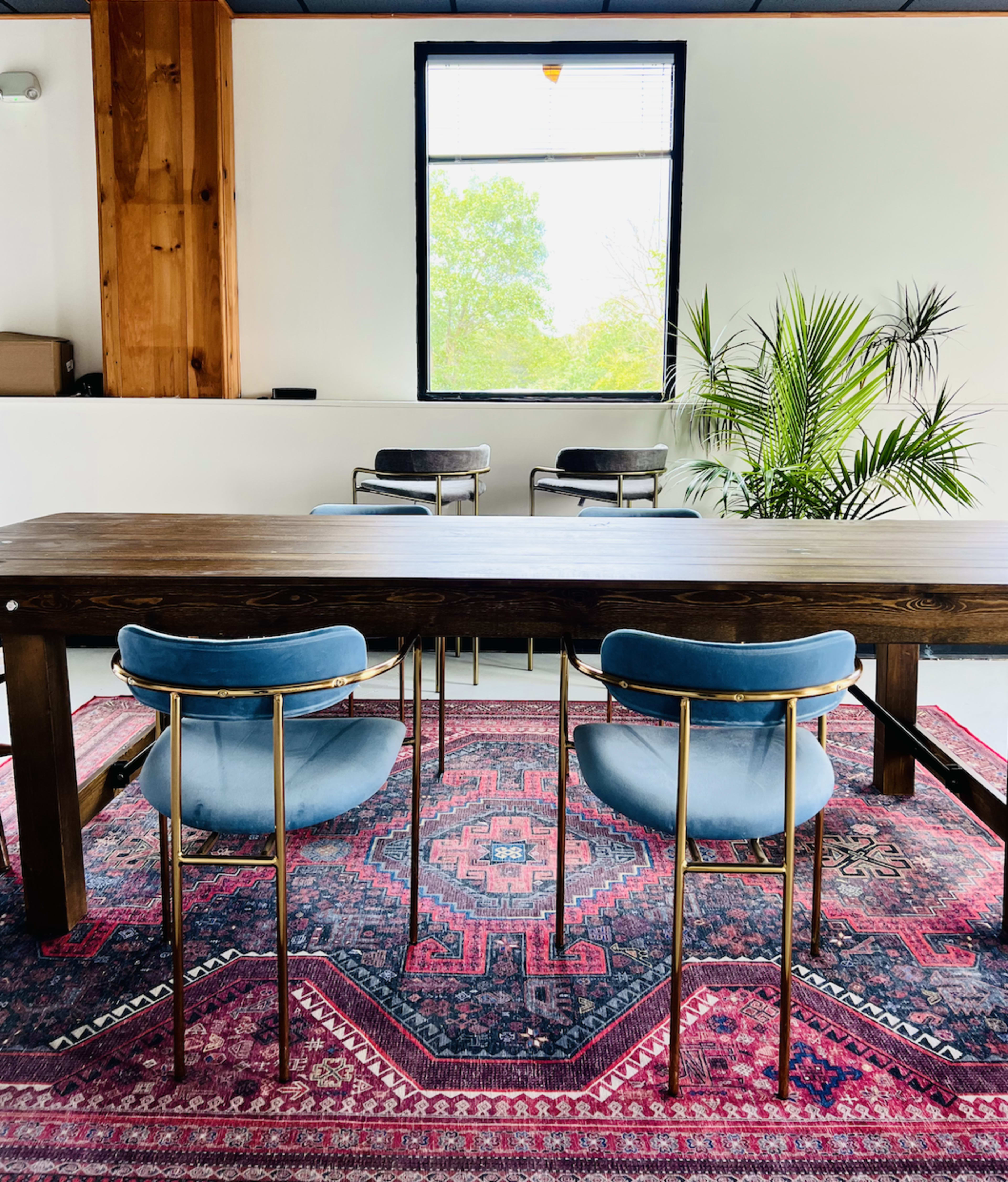 A wooden dining table with two blue velvet chairs and a colorful rug is situated in a room featuring a large window and indoor plants.