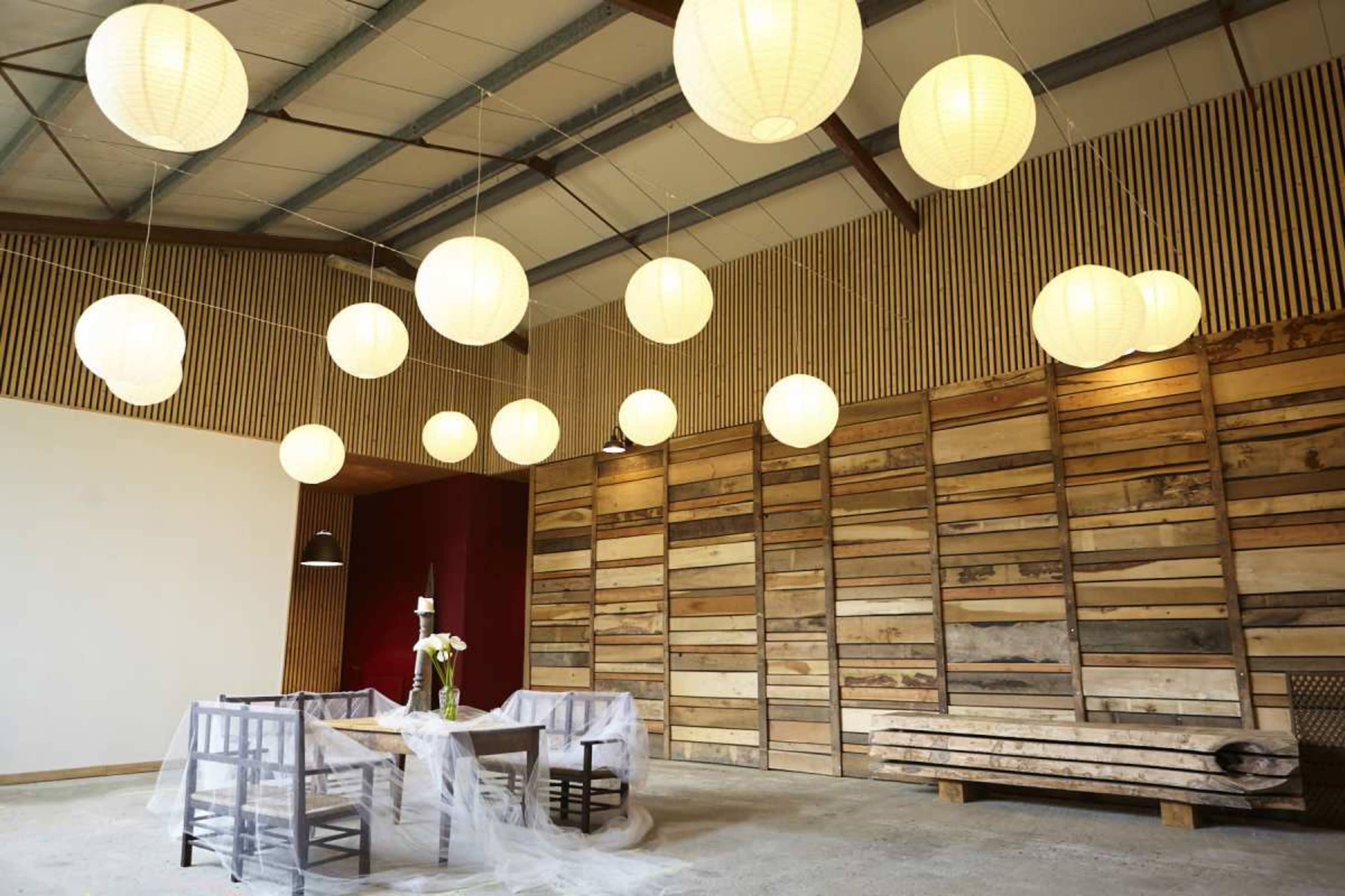 A spacious indoor room with wooden walls, a table set for dining, and multiple hanging lanterns providing light from the ceiling.