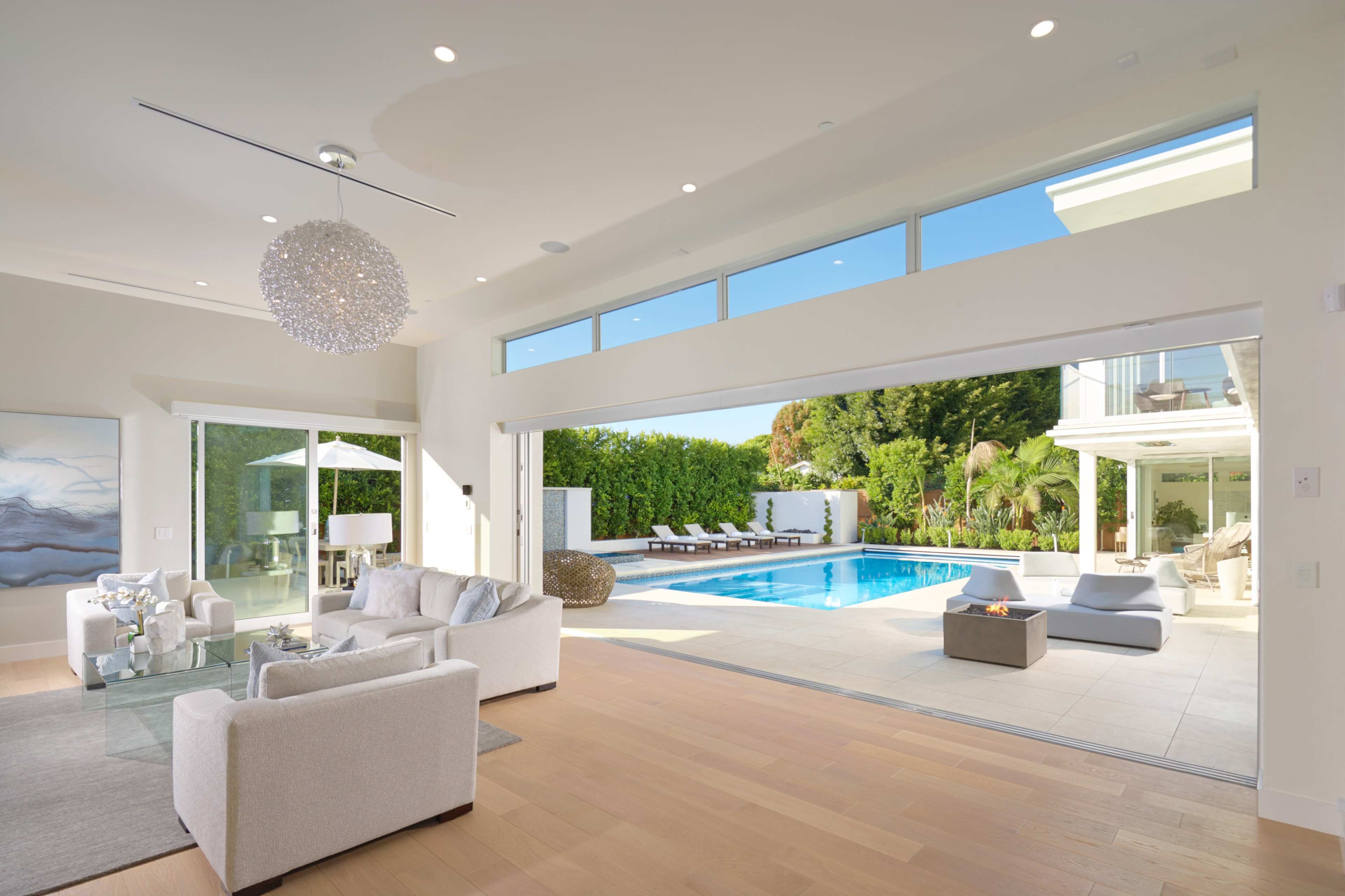 A modern living room opens to a patio and swimming pool surrounded by lush greenery.