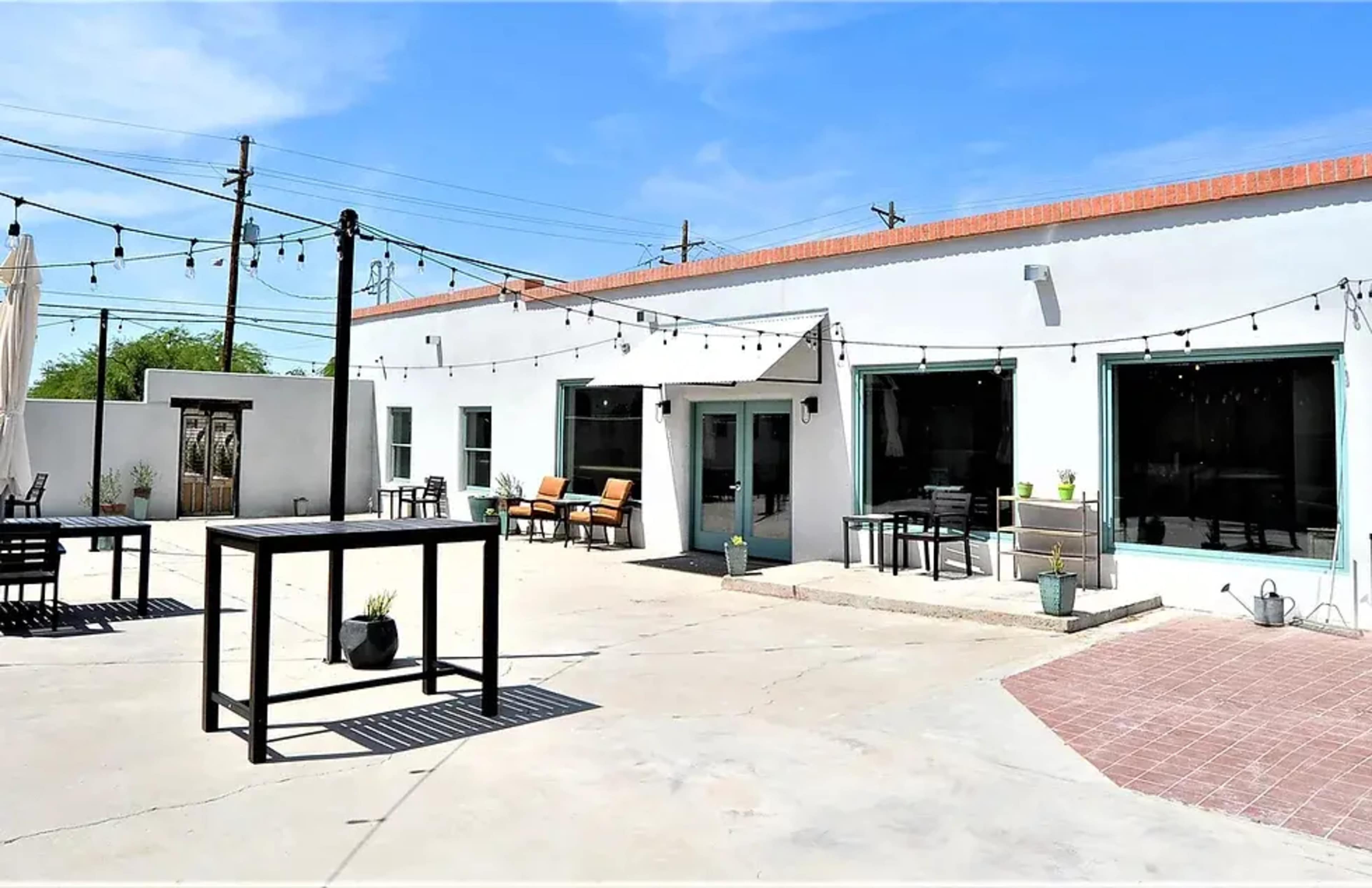 The image shows an outdoor patio area of a modern building with several tables, chairs, and string lights against a clear blue sky.
