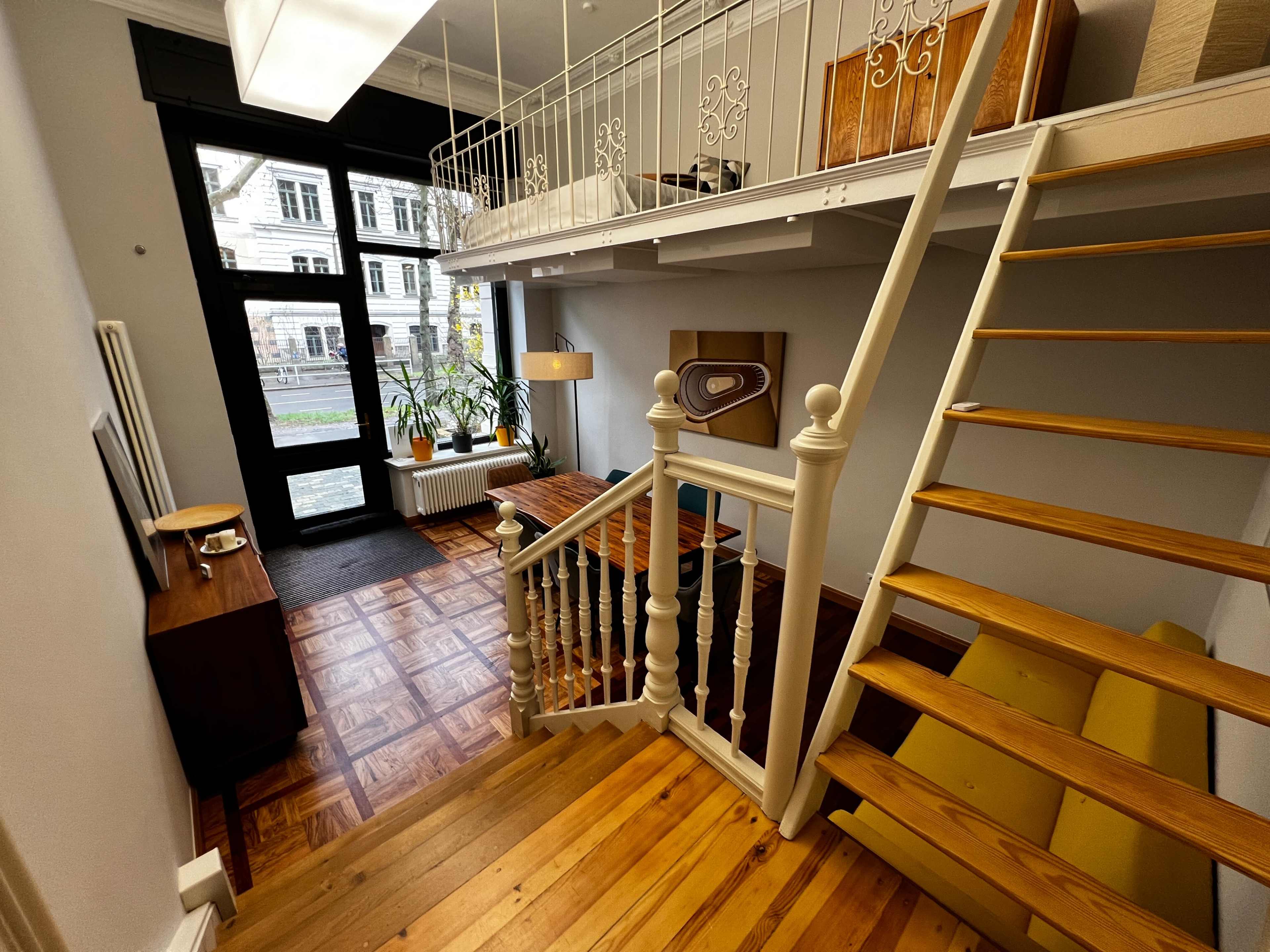 The image shows a cozy interior space with a staircase leading to an upper level, large windows allowing natural light, and a blend of wooden furniture and decorative elements.