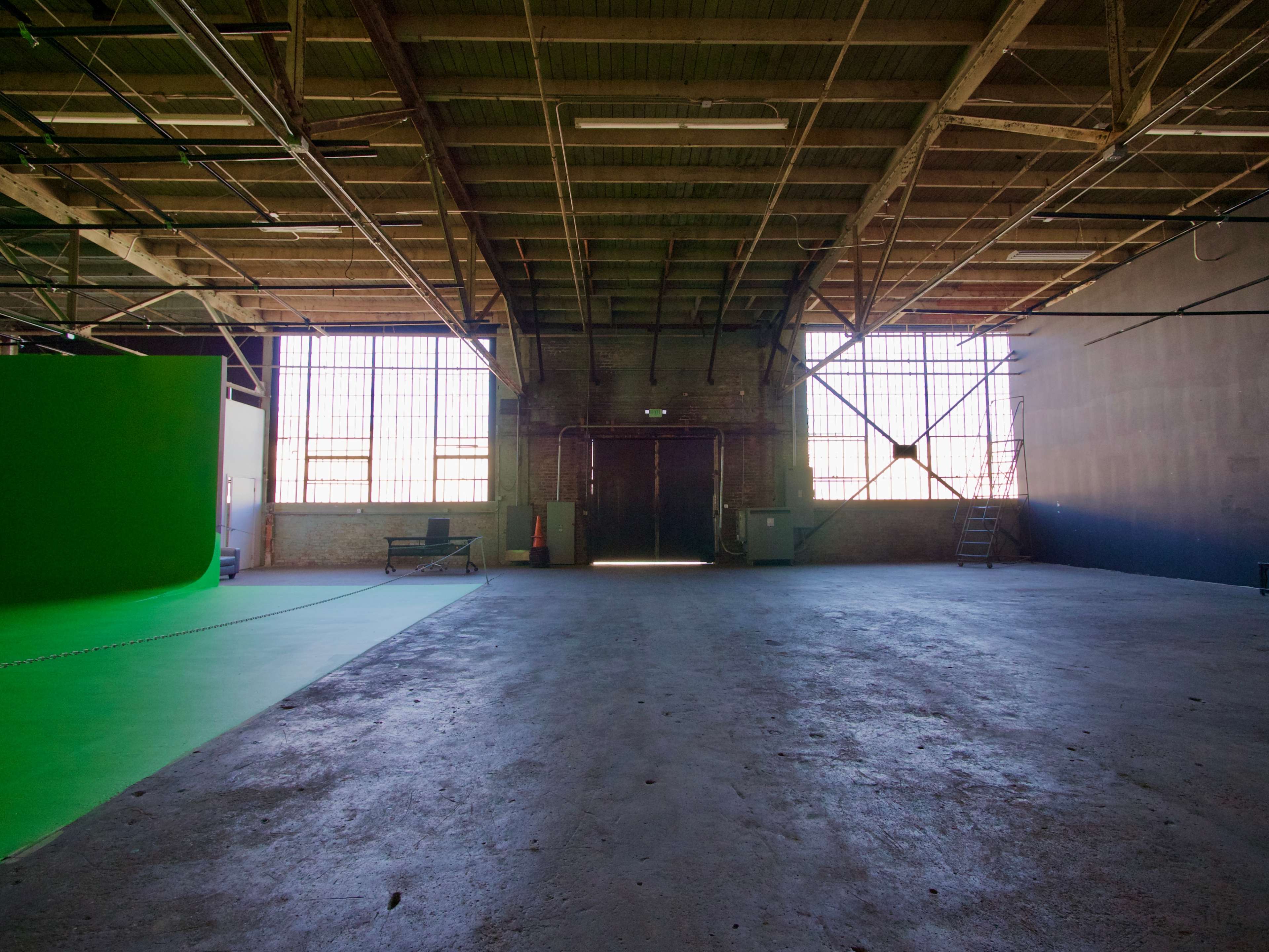 The image shows a large, empty industrial space with green screen setups and large windows allowing natural light.