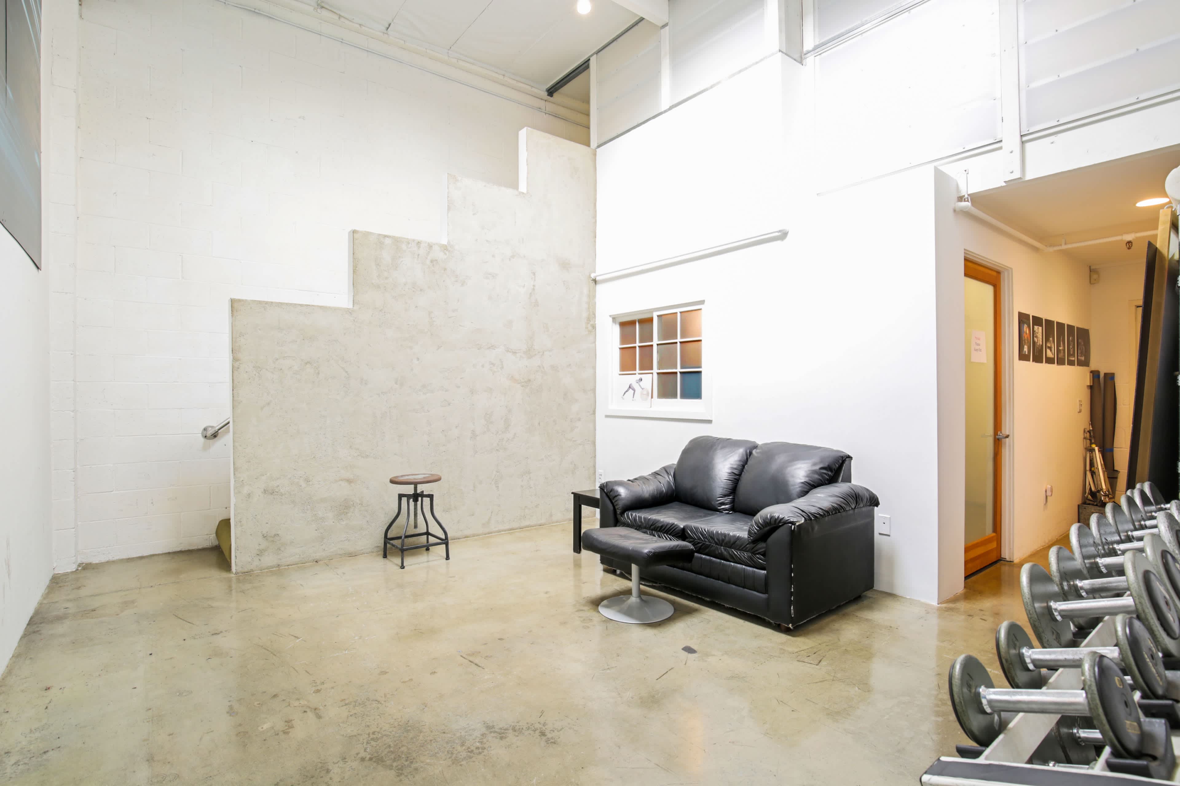 A minimalist gym space featuring a black sofa and a small table next to a set of dumbbells, with a concrete wall and staircase in the background.