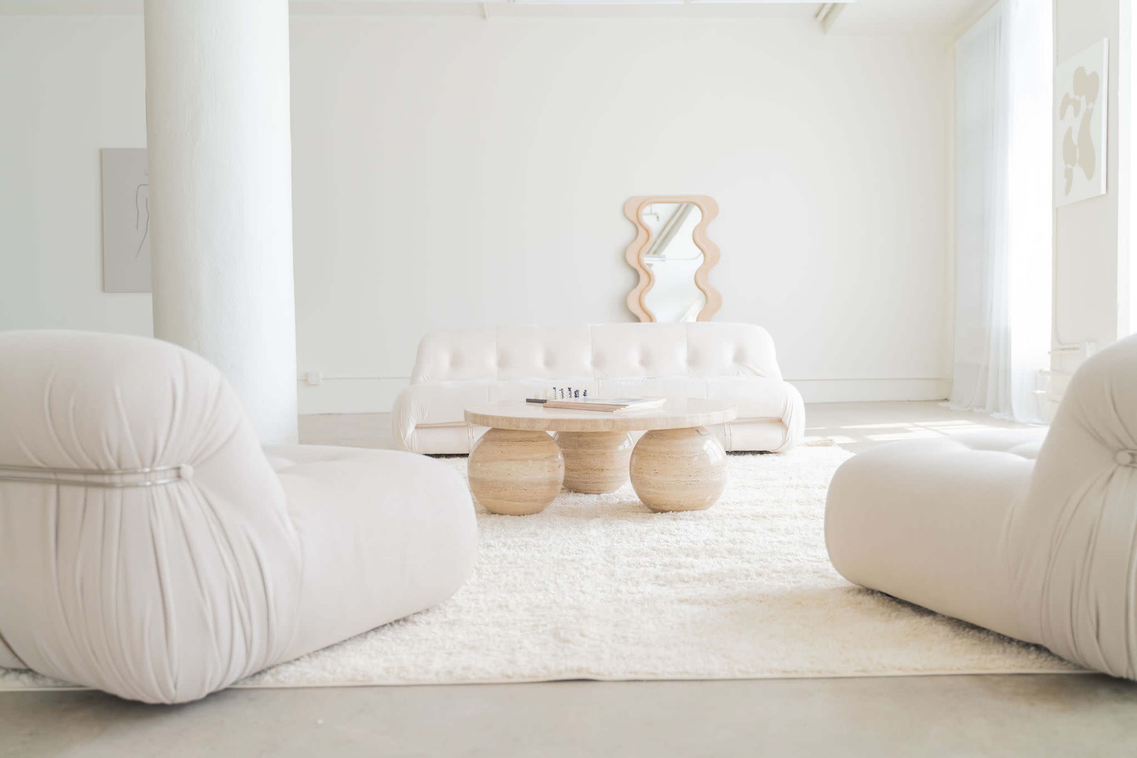 A modern, minimalist living room with a white sofa, two circular wooden coffee tables, and plush armchairs arranged around a light-colored rug.