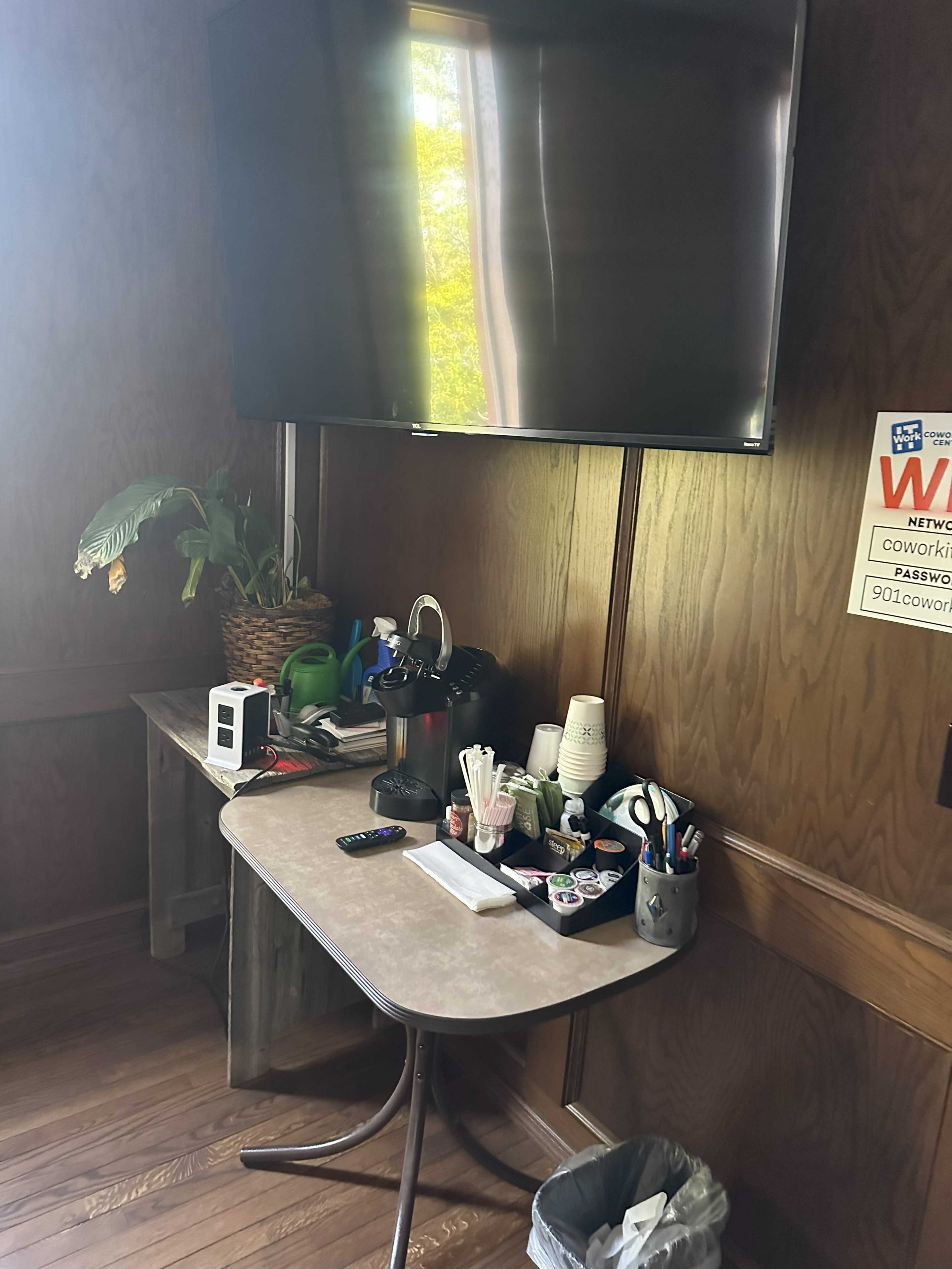 A small table is set up against a wooden wall, displaying a coffee maker, cups, and various office supplies, with a flat-screen TV mounted above it.