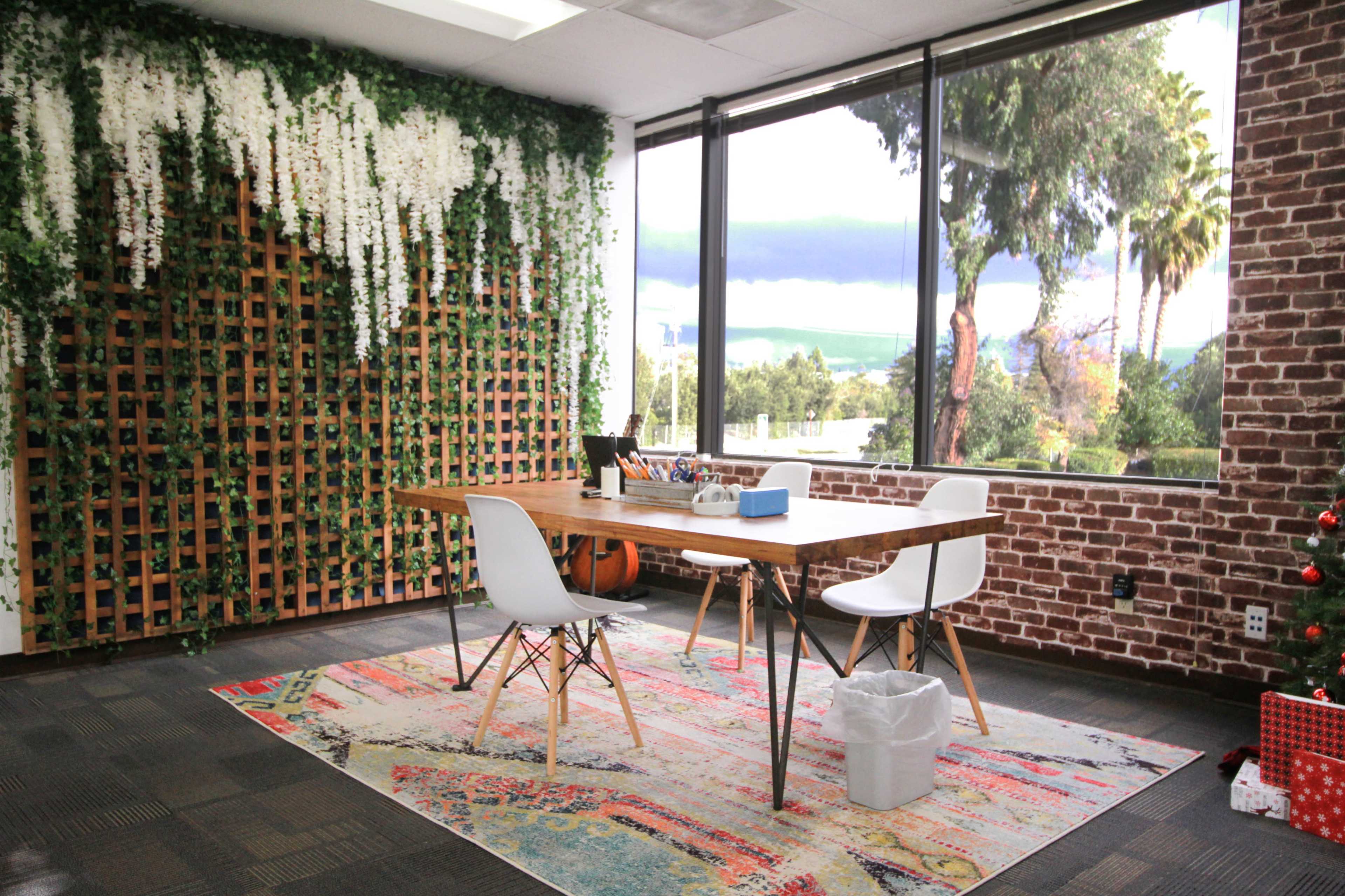 A modern office space features a wooden table surrounded by white chairs, set against a backdrop of greenery and large windows with an outdoor view.