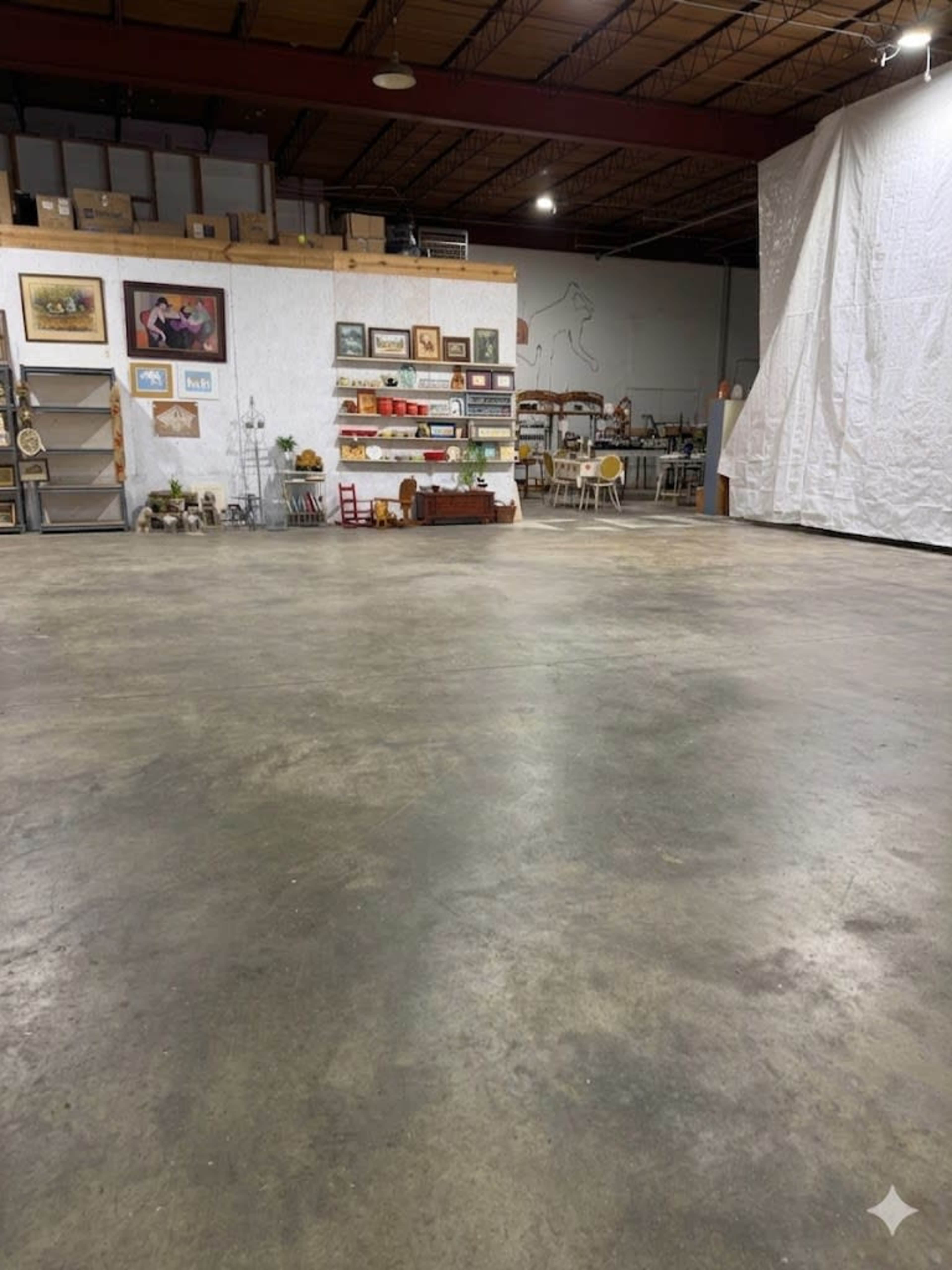 The image shows an empty warehouse space with a concrete floor, featuring shelves adorned with various items and a large white curtain on one side.