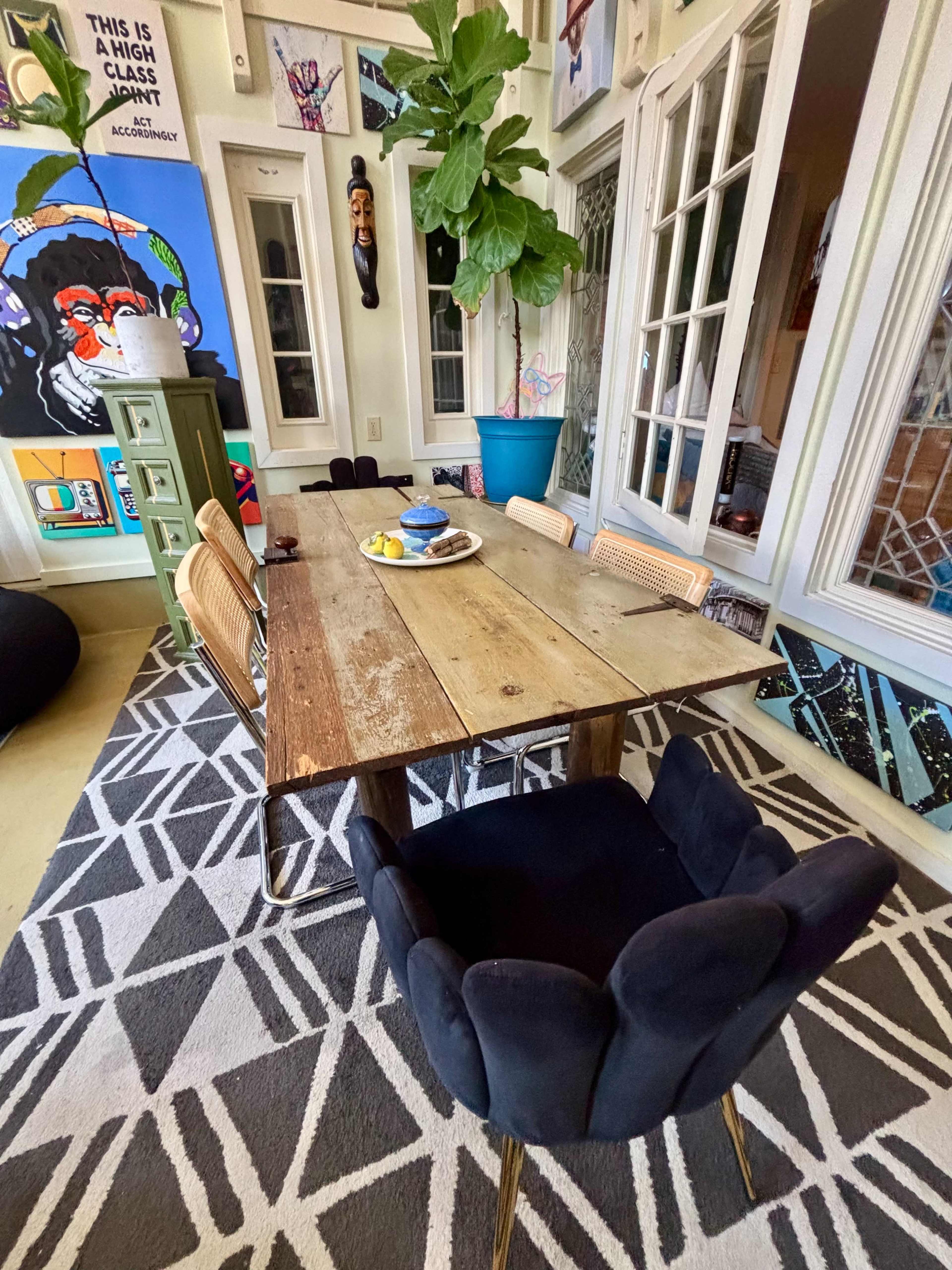 A rustic wooden dining table is set in a brightly decorated room, surrounded by various chairs and a large potted plant.