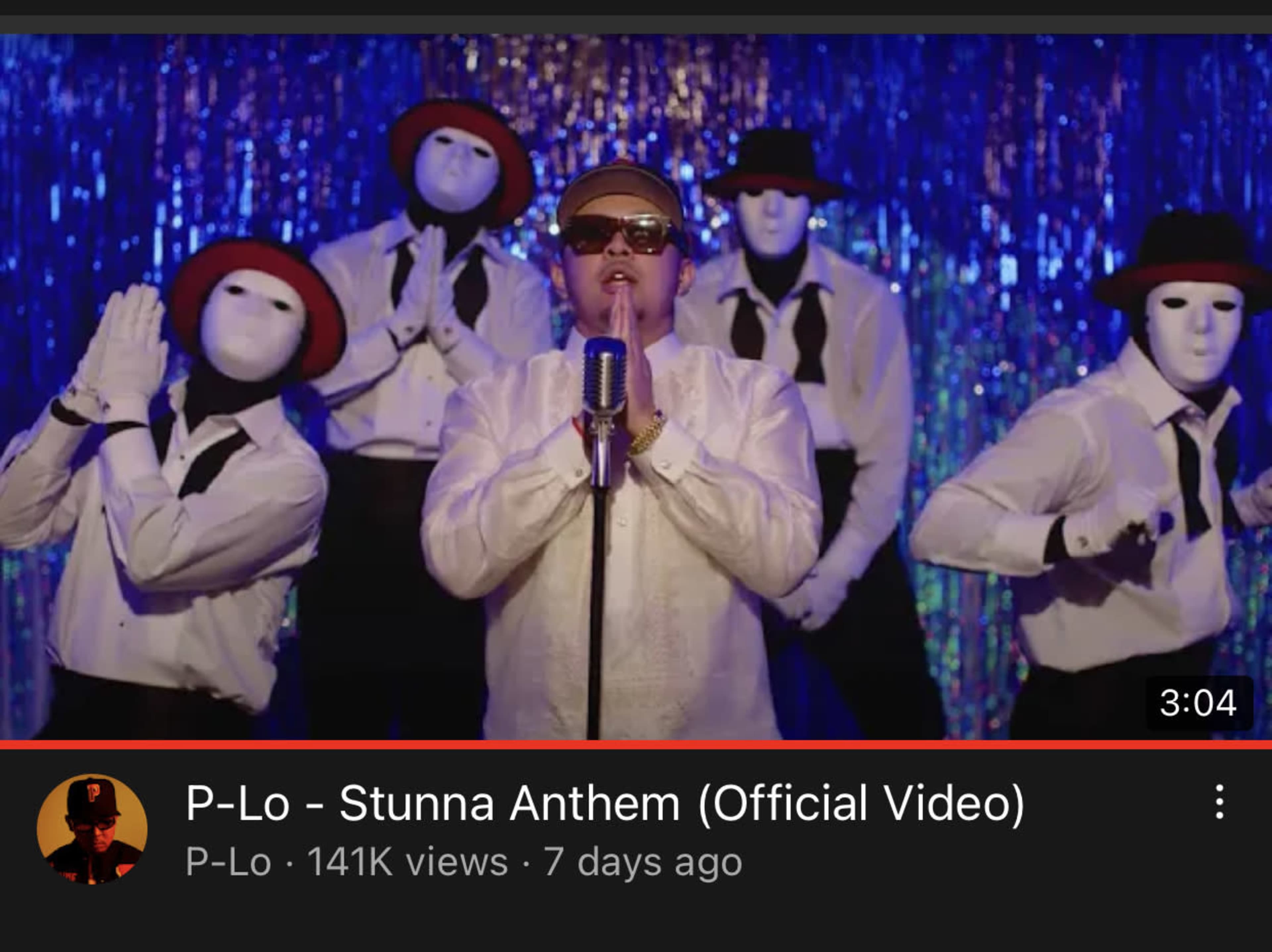 A male performer stands at a microphone with five dancers in suits and white masks posed behind him against a glittering backdrop.