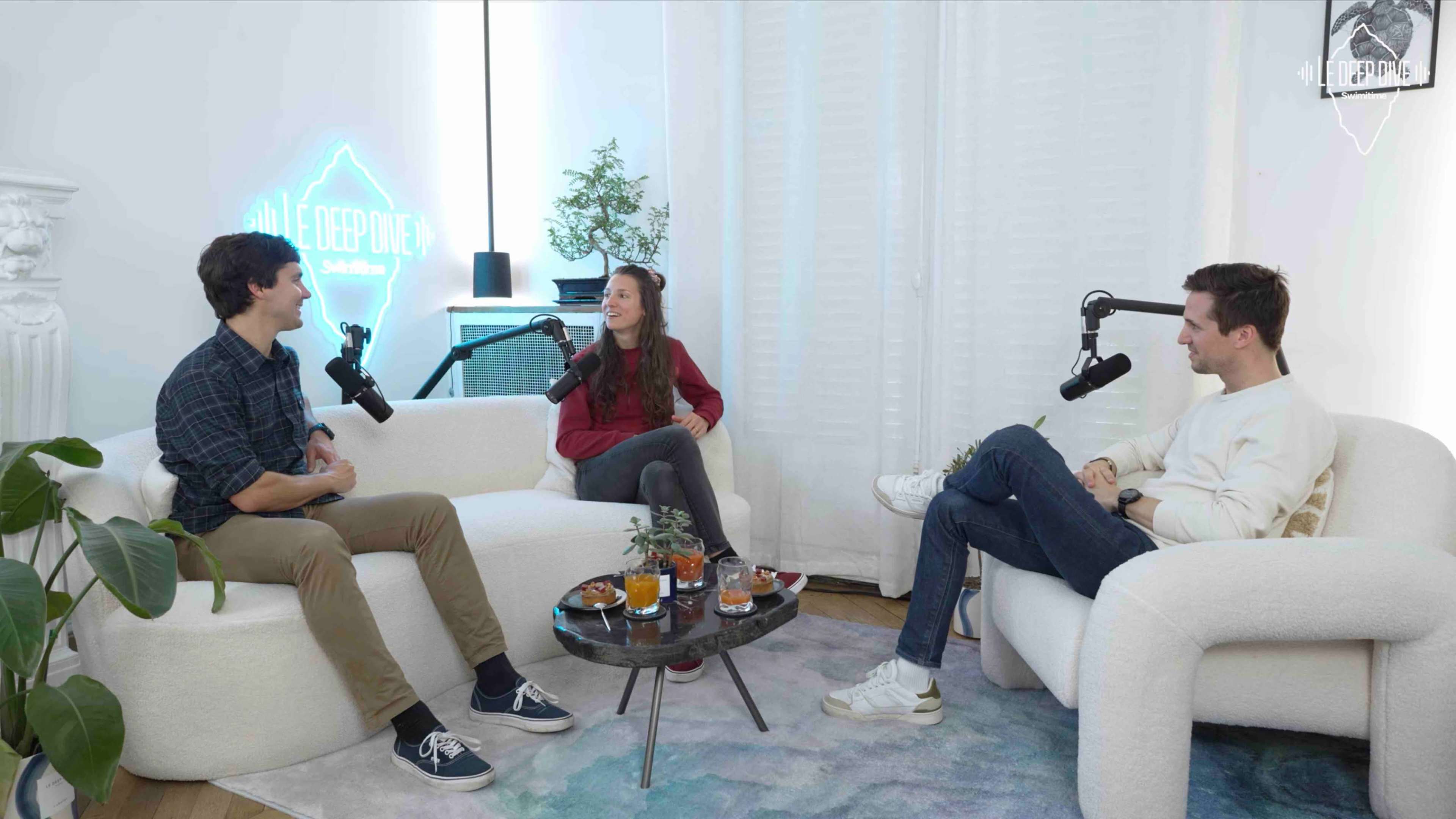 Three people sit in a cozy, modern studio with microphones, discussing at a circular coffee table adorned with small snacks and drinks.