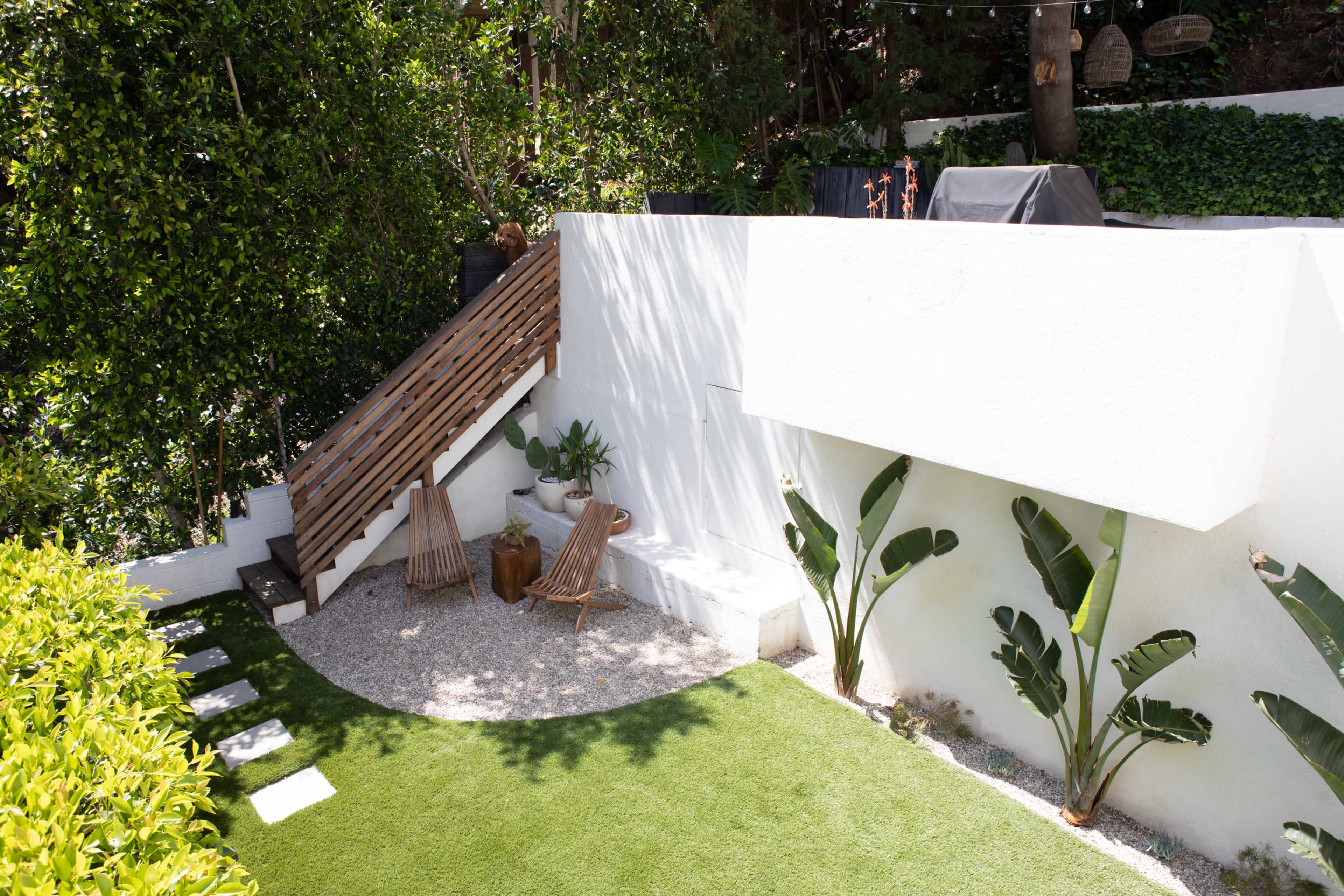A landscaped garden area with a staircase leading to a raised platform, surrounded by plants and featuring two wooden chairs on a gravel surface.