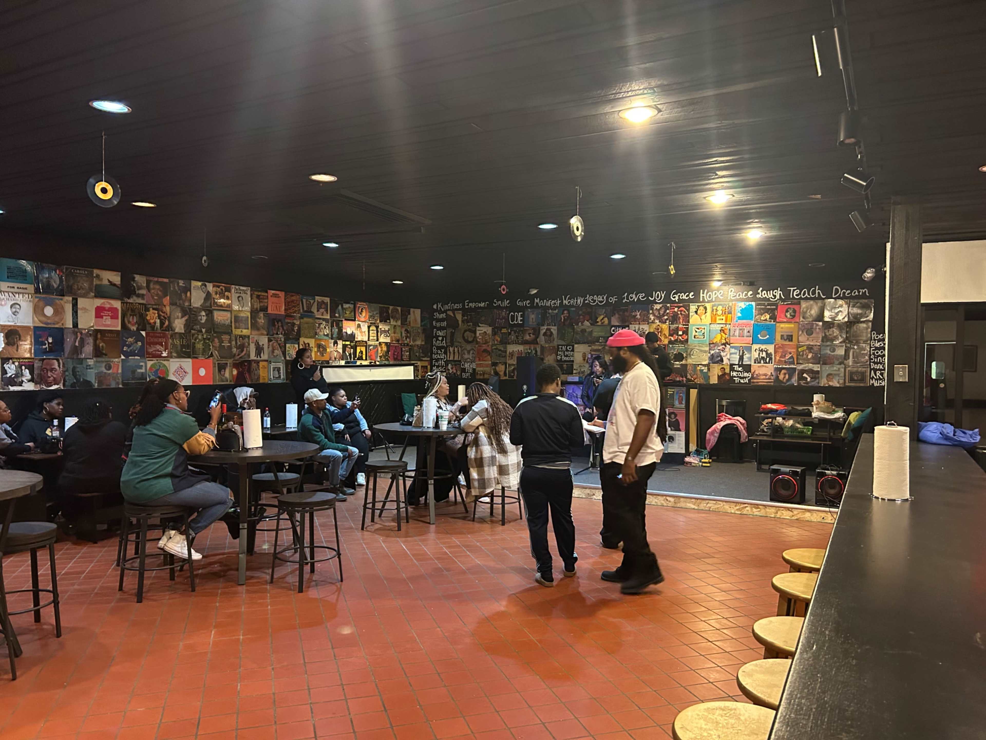 A dimly lit bar or lounge area with patrons seated at small tables, surrounded by walls decorated with vinyl records and large murals.