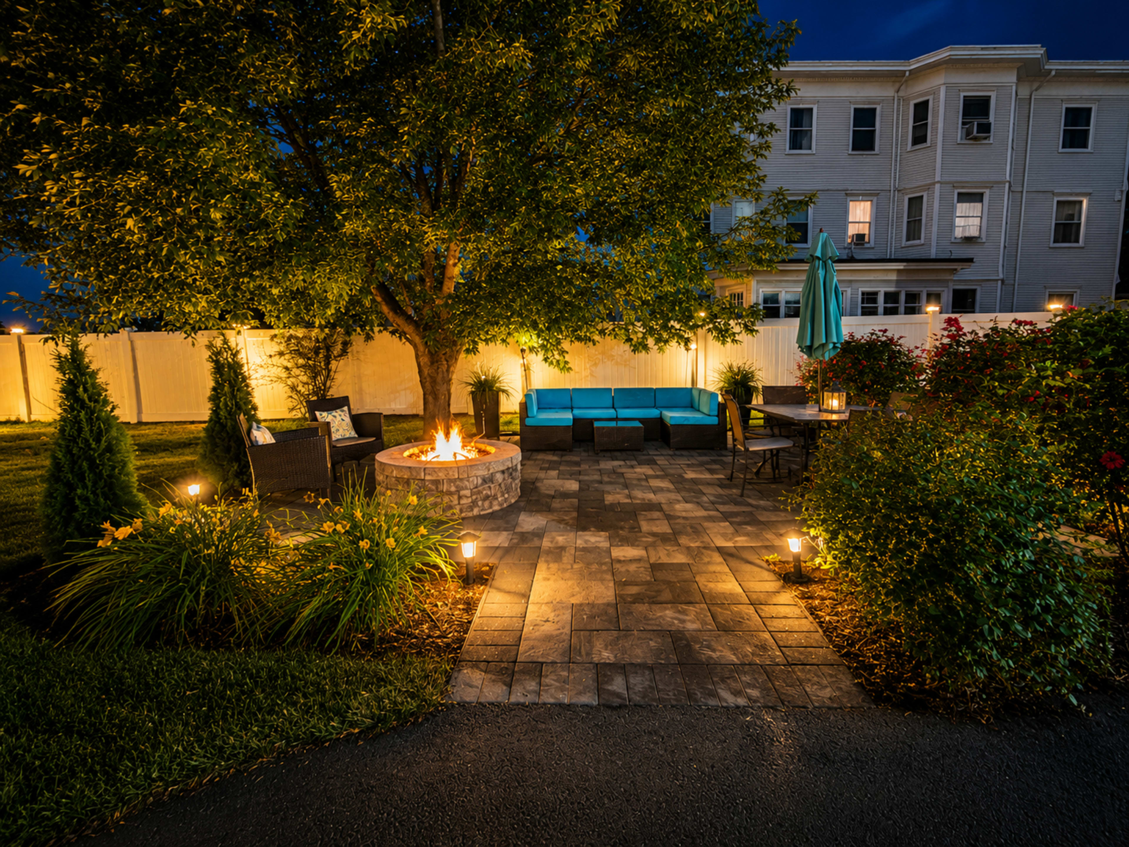 The image shows a landscaped backyard with a stone fire pit surrounded by seating, illuminated by pathway lights and featuring a large tree.