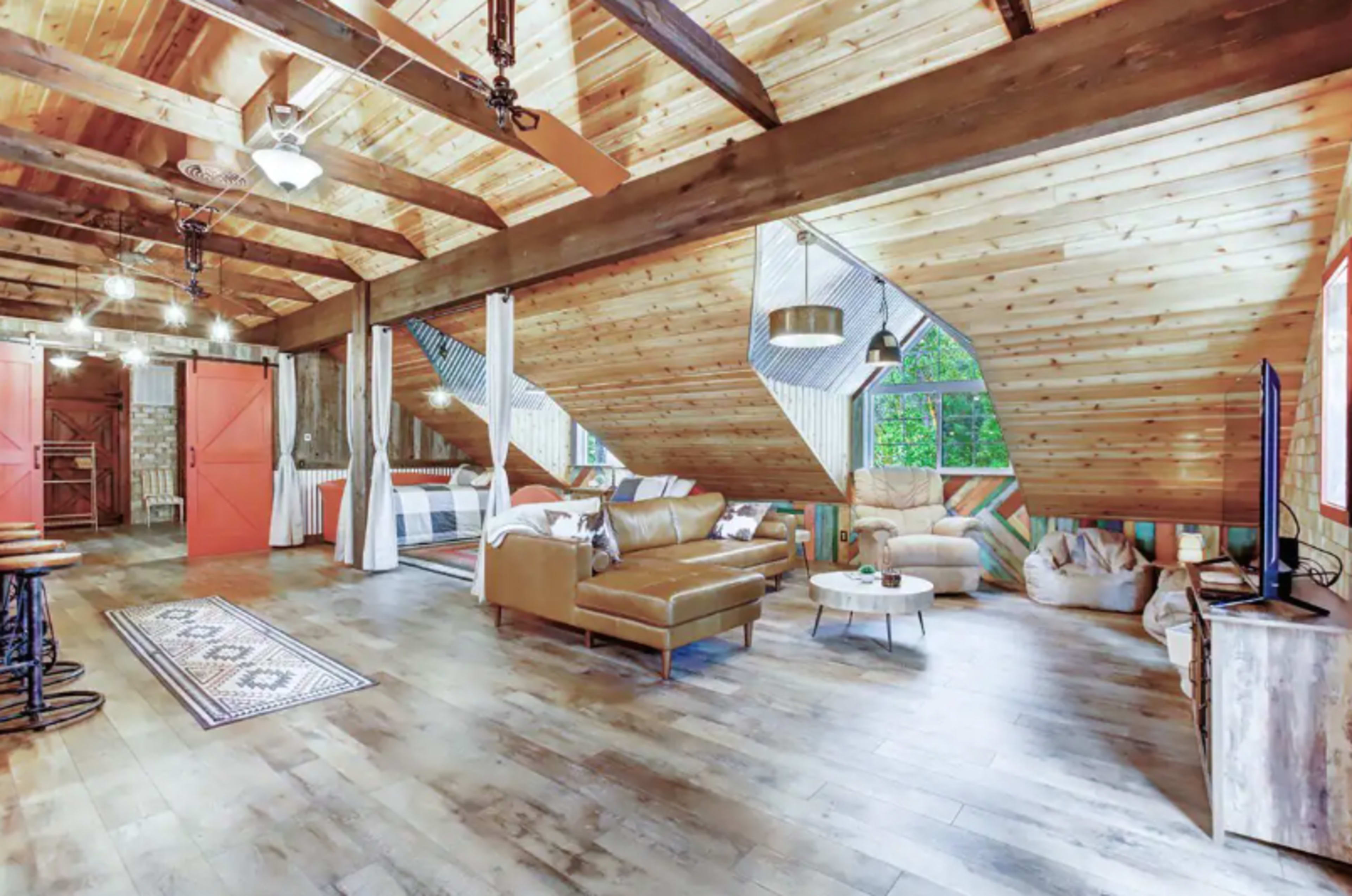 The room features a spacious, rustic interior with wooden beams, a leather sofa, and large windows that allow natural light to fill the space.