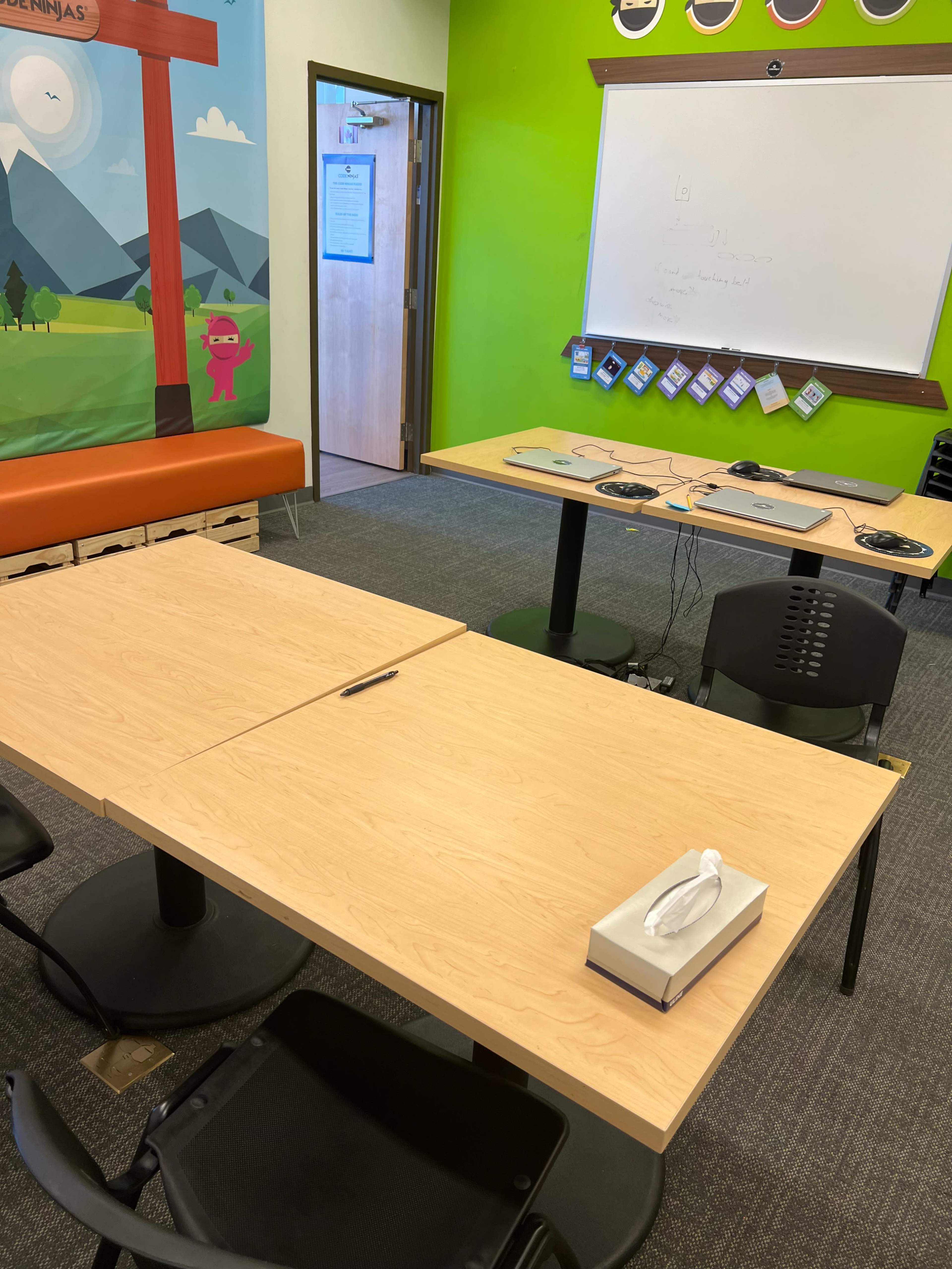 A small meeting room with two wooden tables, black chairs, and some electronic equipment, including laptops and a tissue box, against a colorful mural on one wall.