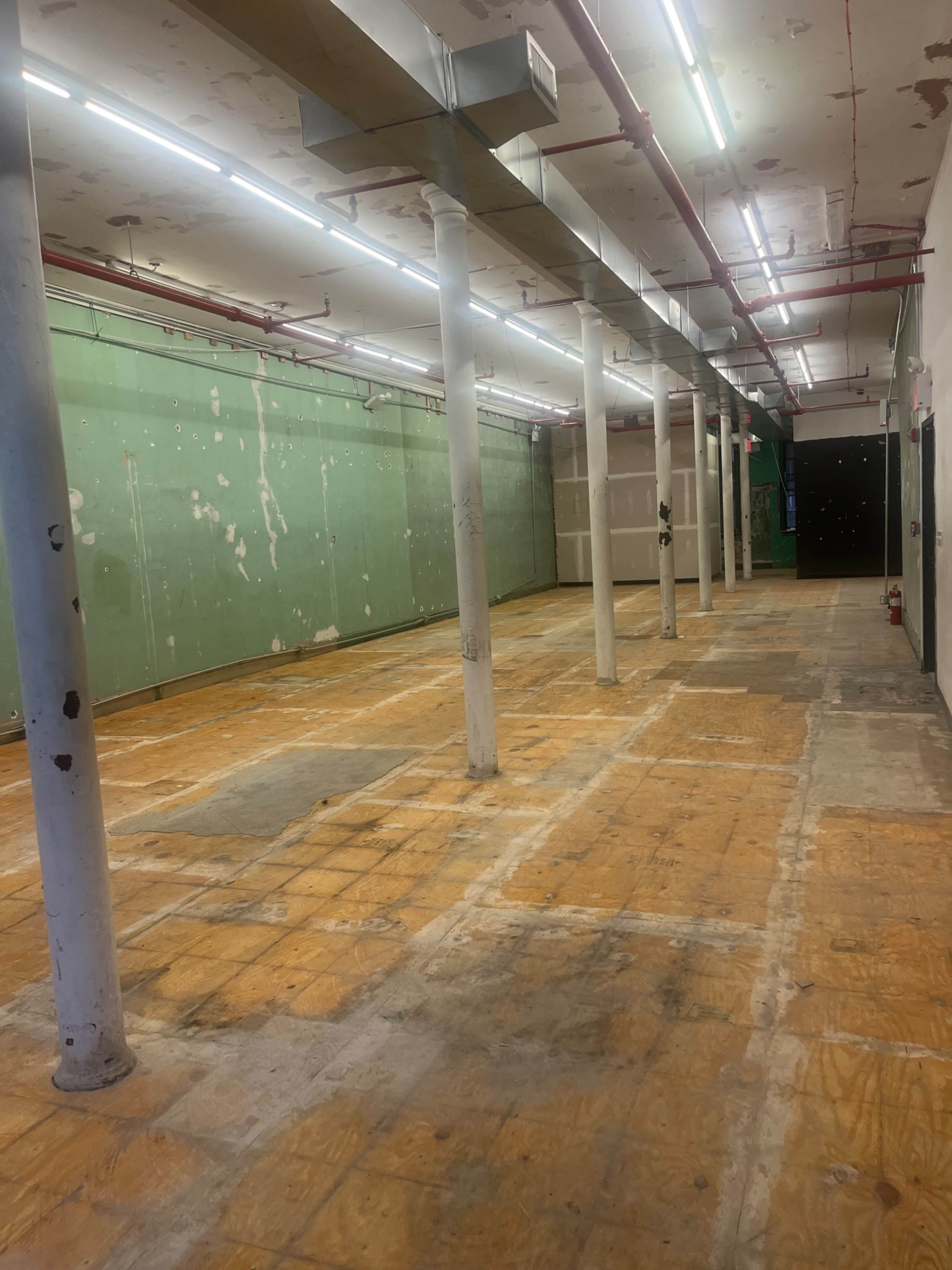 The image shows a large, empty commercial space with wooden flooring and several concrete columns, illuminated by fluorescent lights.