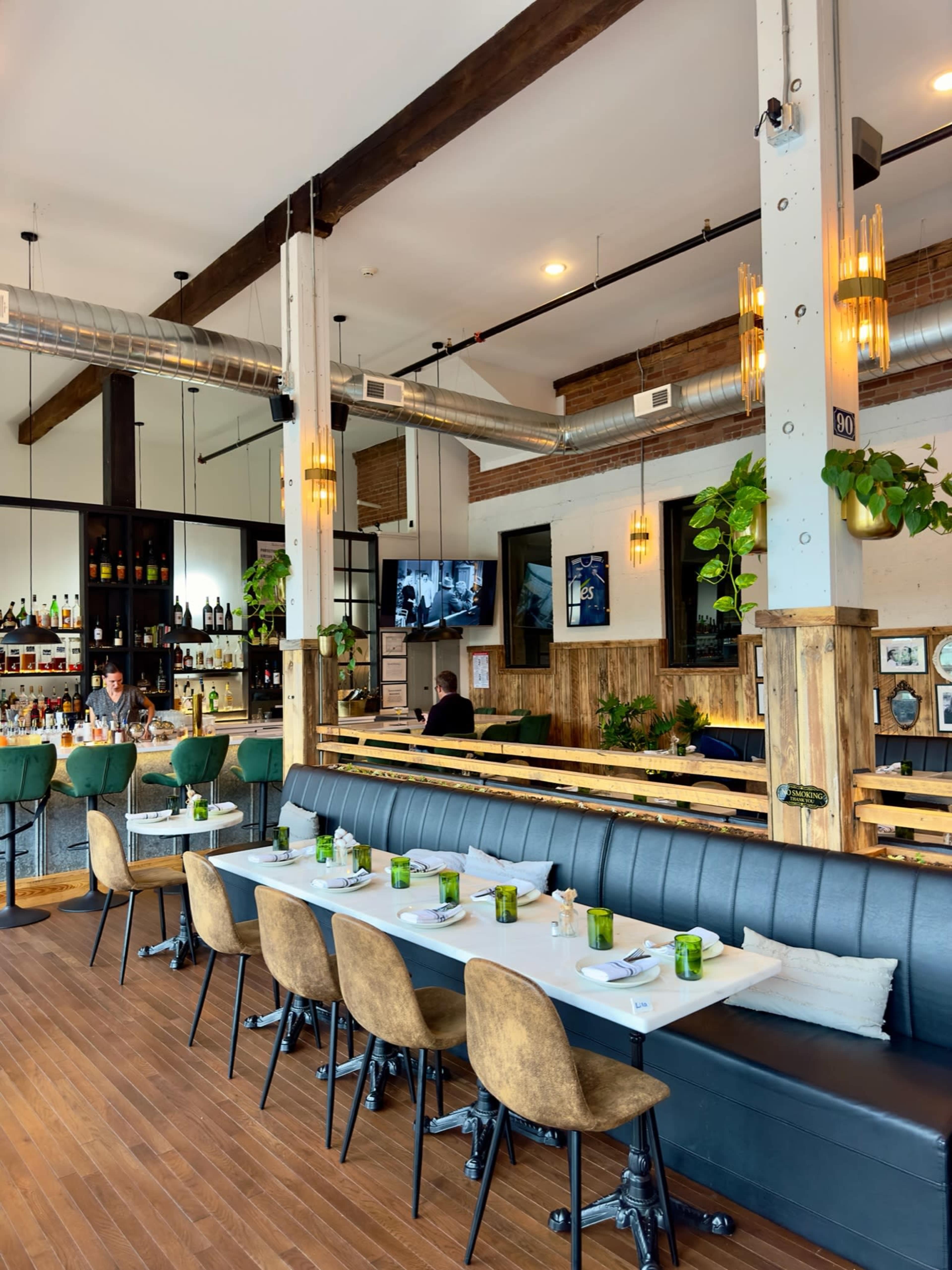 The image shows a modern restaurant interior with a wooden floor, leather seating, and a bar area in the background.