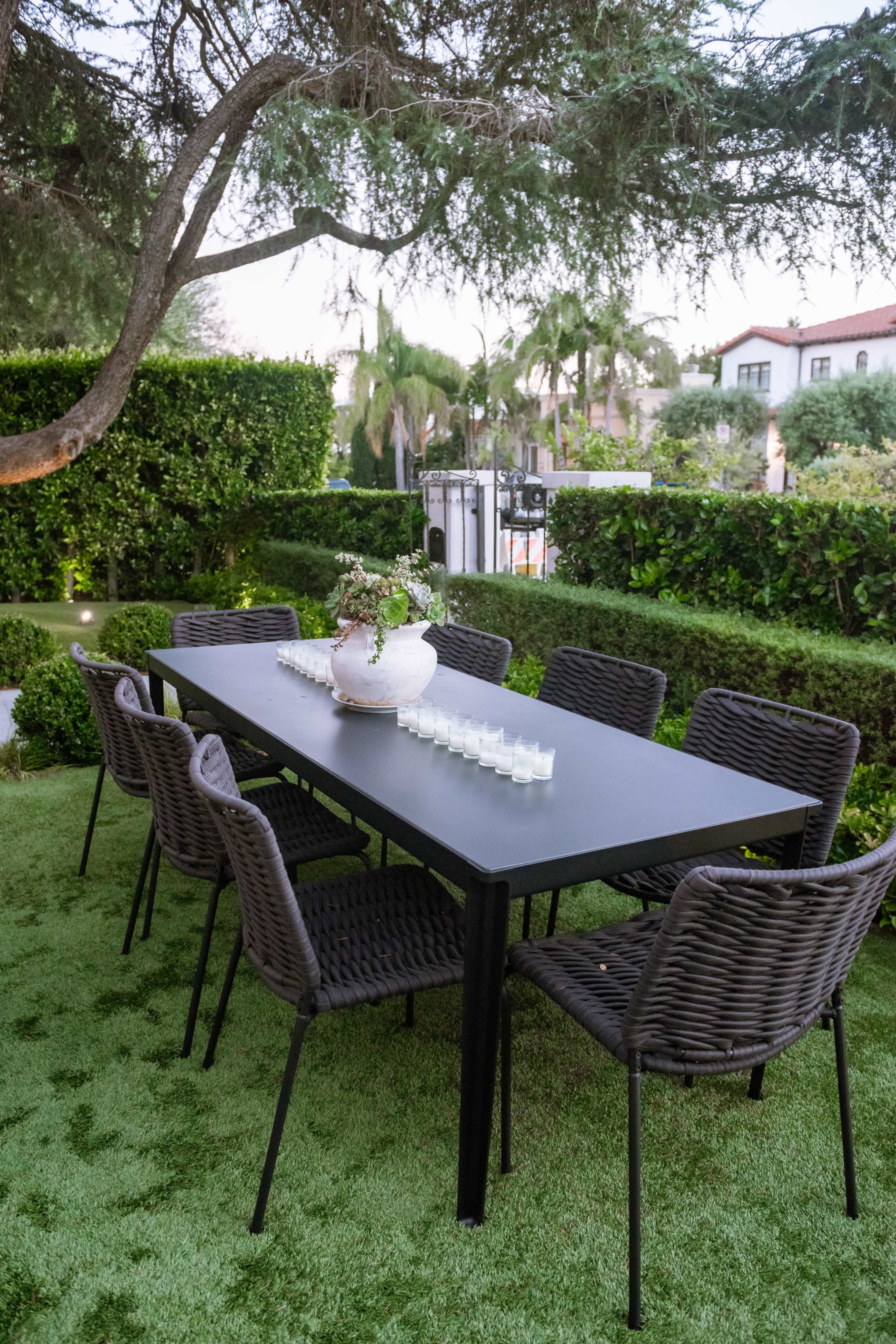 A black dining table surrounded by eight chairs is set up on a grassy area, with a decorative vase in the center and hedges and trees in the background.