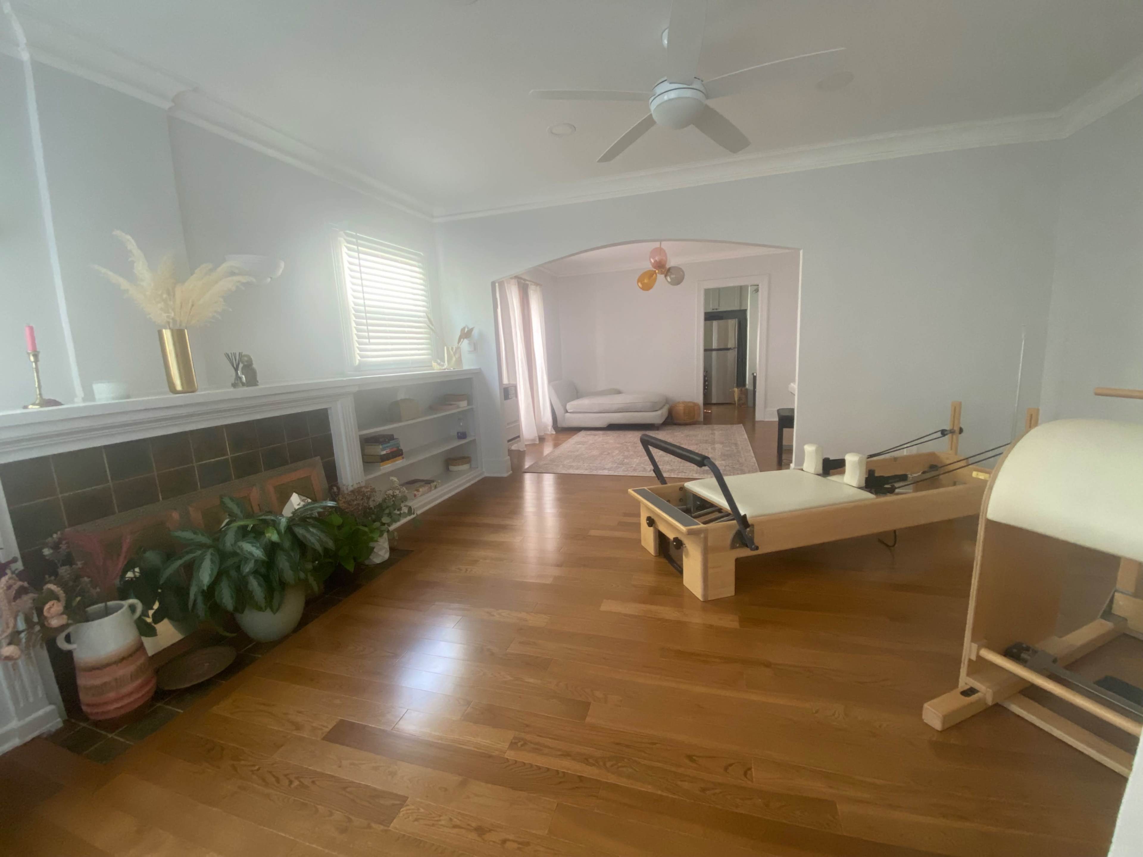 The image shows a bright, open living space with a wooden floor, a pilates reformer, plants, and a cozy seating area in the background.