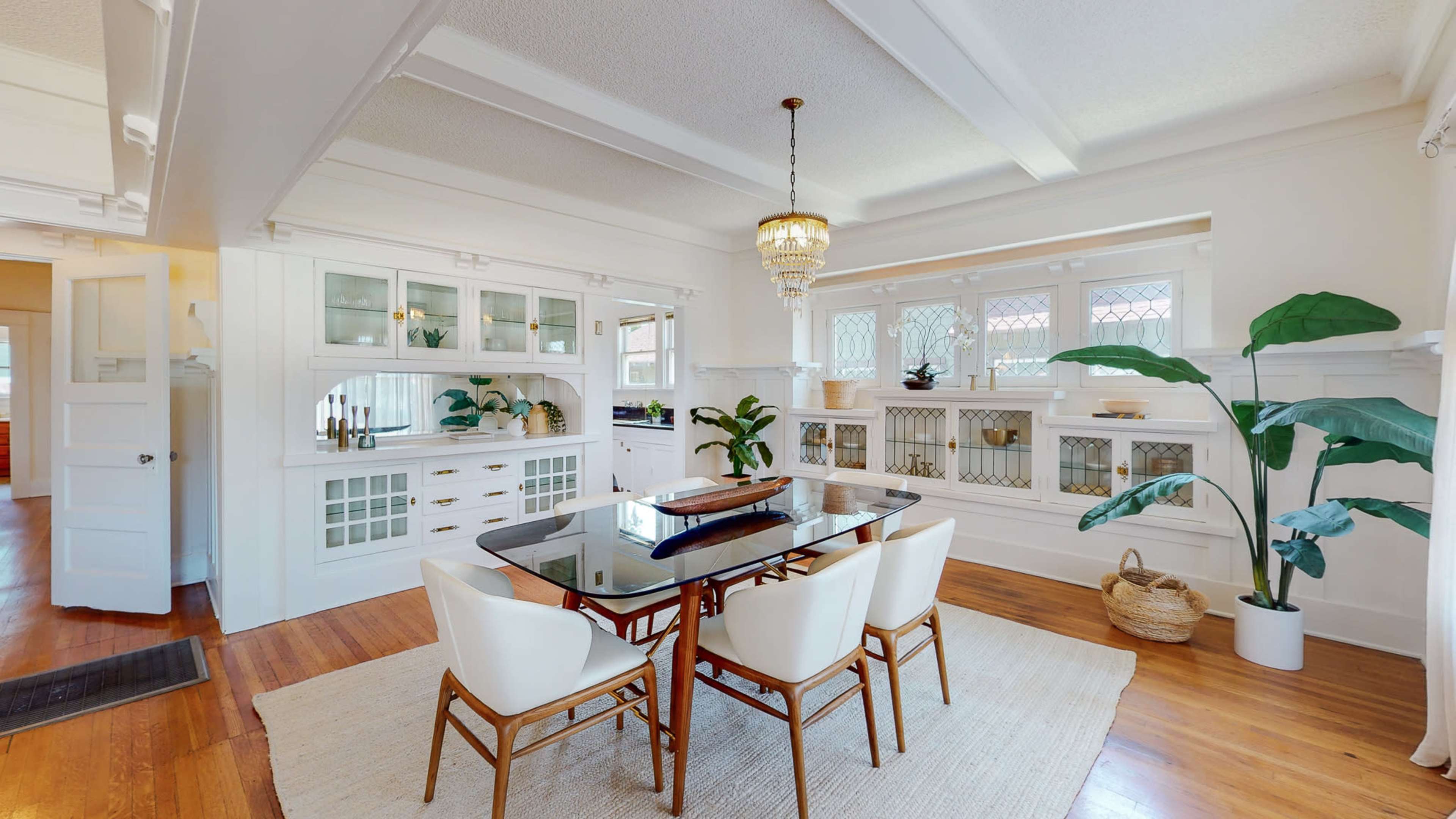 A dining area features a glass-top table surrounded by white chairs, with built-in cabinetry and large windows displaying plants.