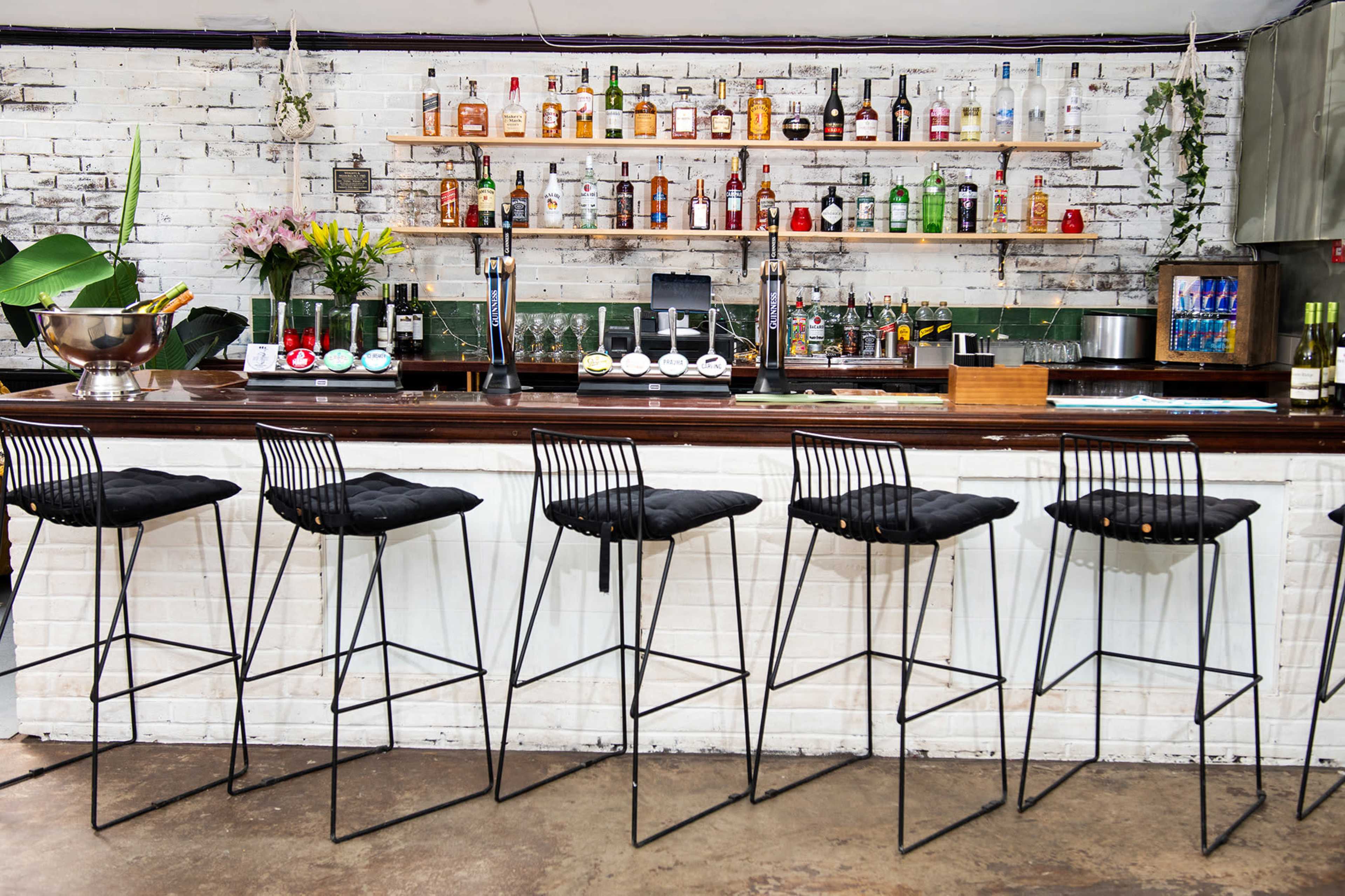 A modern bar with a white brick wall displays a variety of liquor bottles on shelves above a dark wooden counter and black wire bar stools.