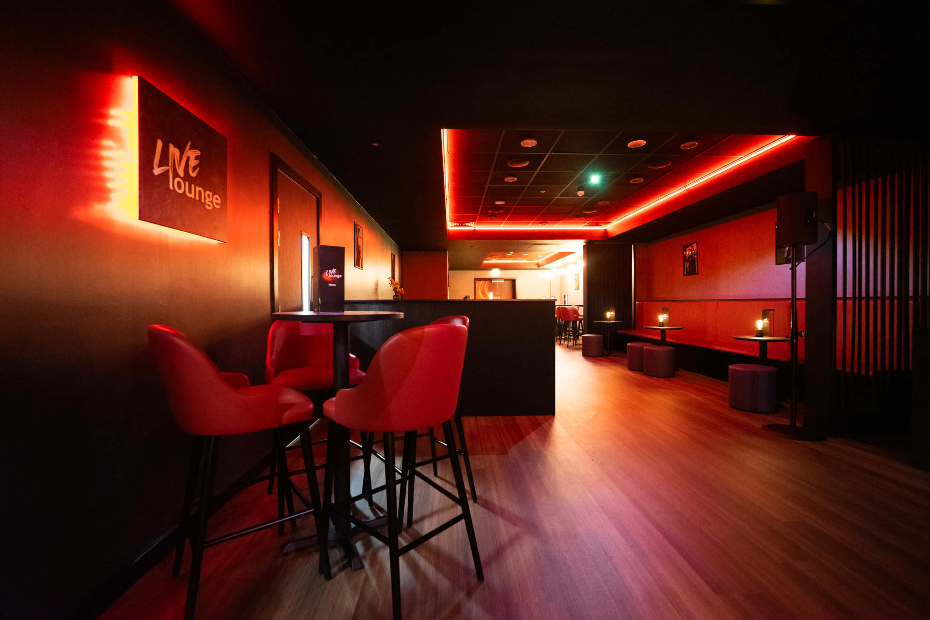 The image shows a modern lounge featuring red lighting, high tables with red chairs, and a sleek, minimalistic design.