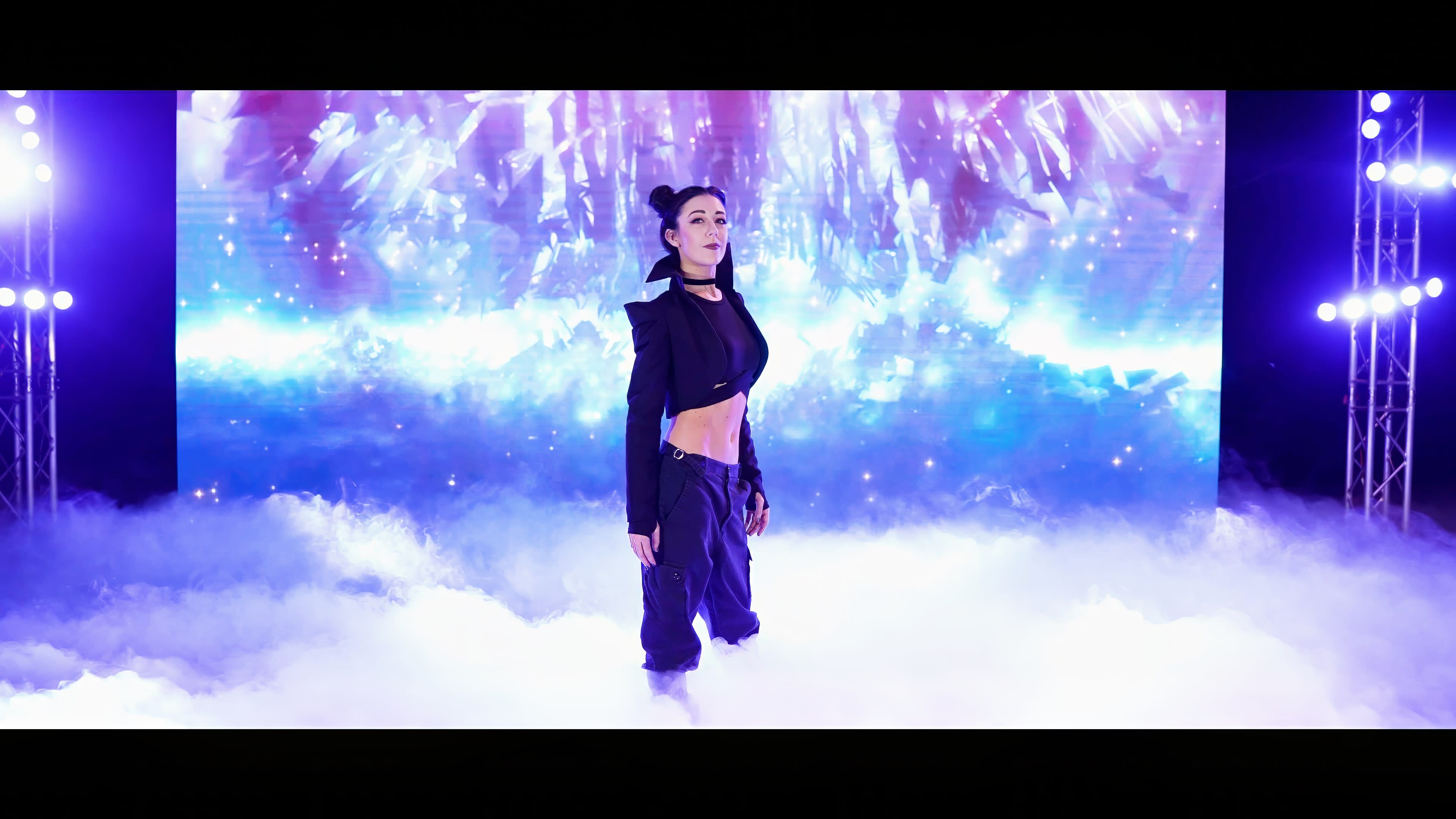 A performer stands confidently in a cloud of mist against a vibrant, illuminated backdrop.