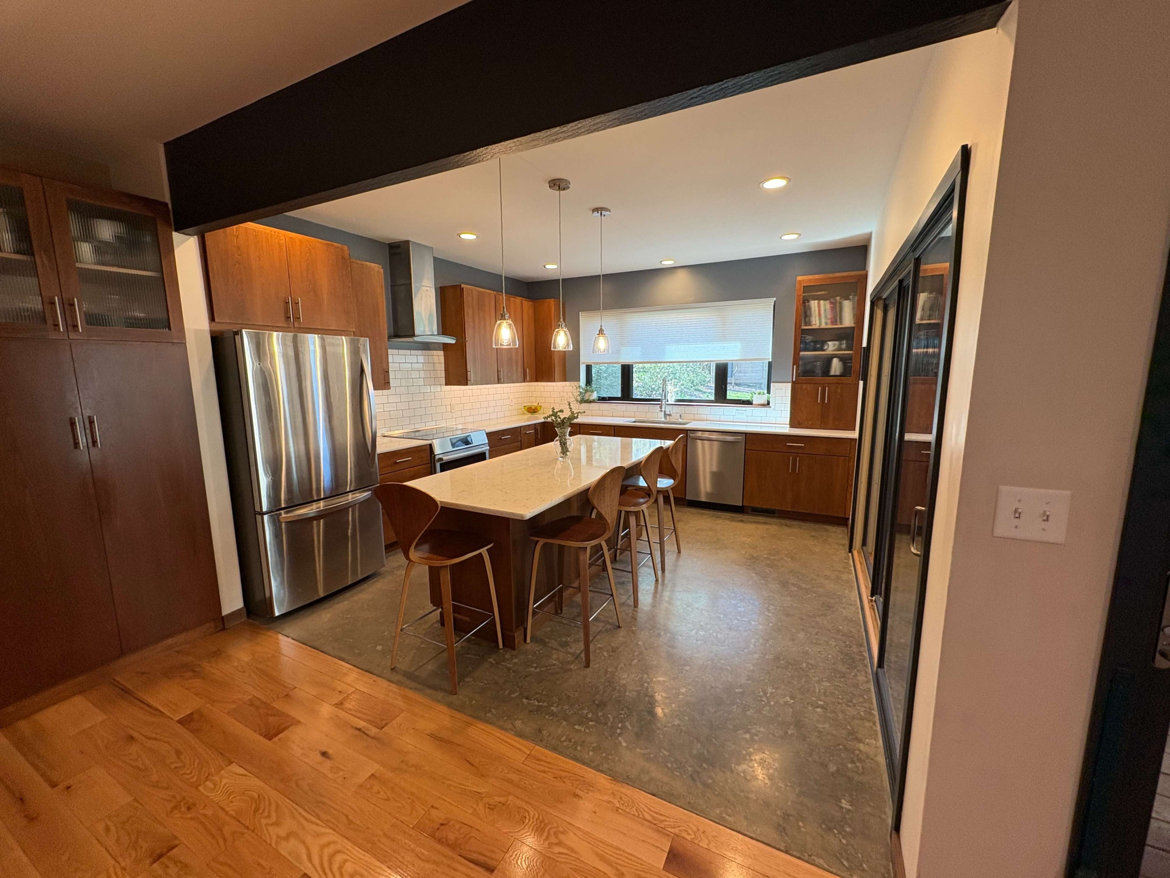 The image shows a modern kitchen with wooden cabinetry, stainless steel appliances, and a central island with seating.