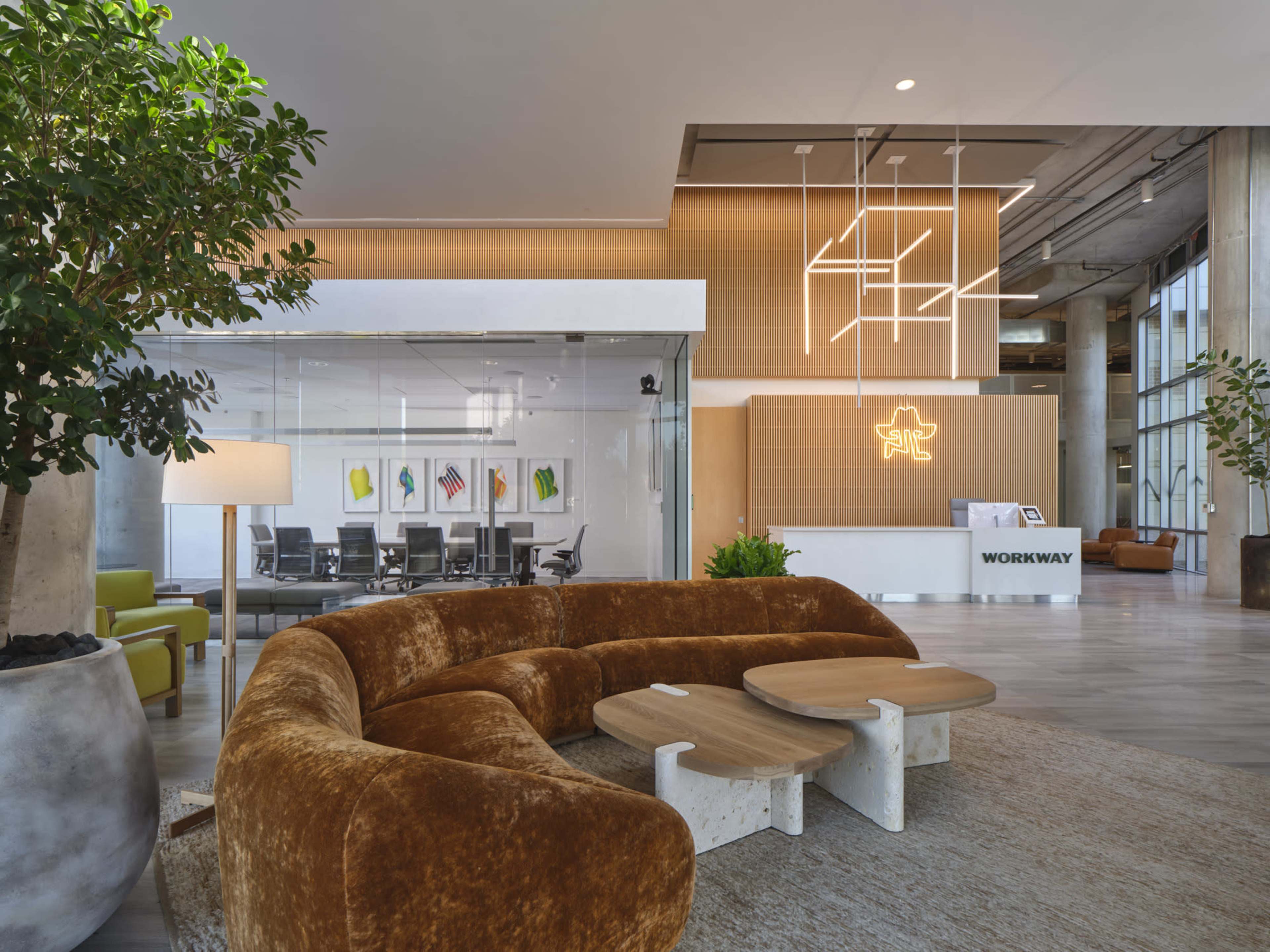 The image shows a modern office lobby featuring a large, brown, curved sofa, a couple of wooden coffee tables, and a glass-walled meeting room in the background with a reception desk displaying a glowing sign.