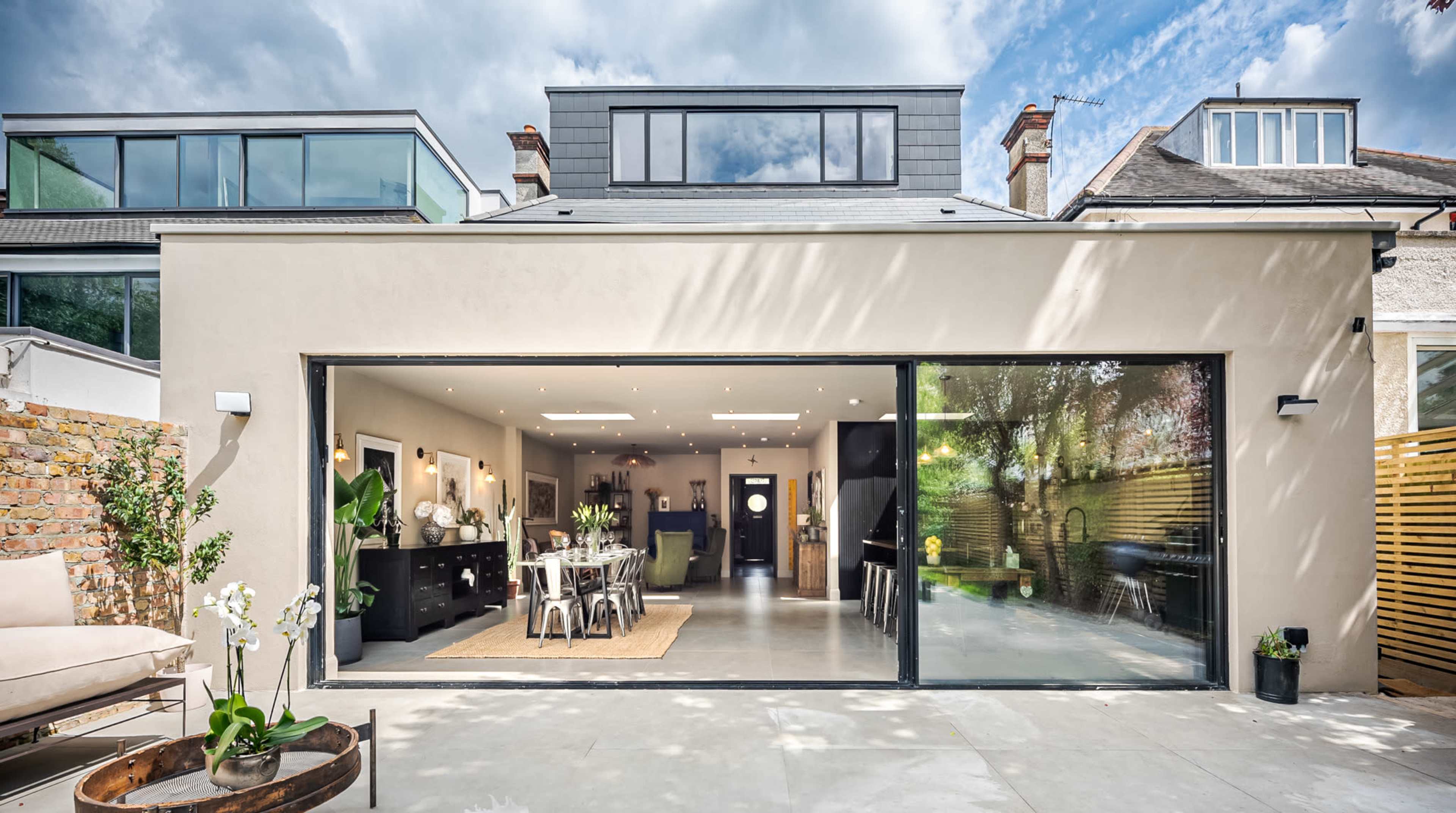 The image shows a modern residential extension with large glass doors opening to a dining area, featuring a contemporary design and a spacious outdoor patio.