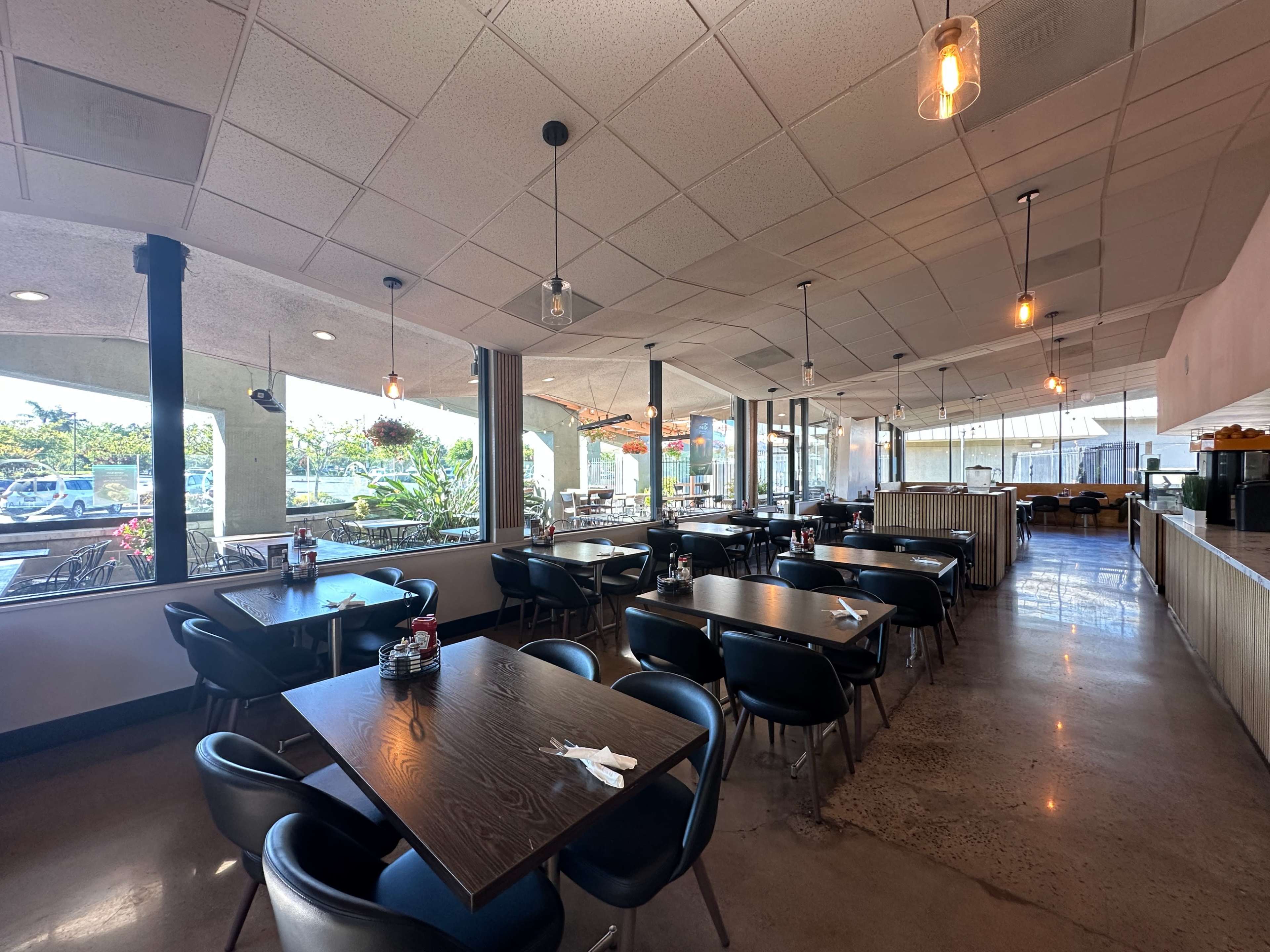 The image shows an interior of a restaurant featuring rows of tables with chairs, large windows letting in natural light, and a modern decor.