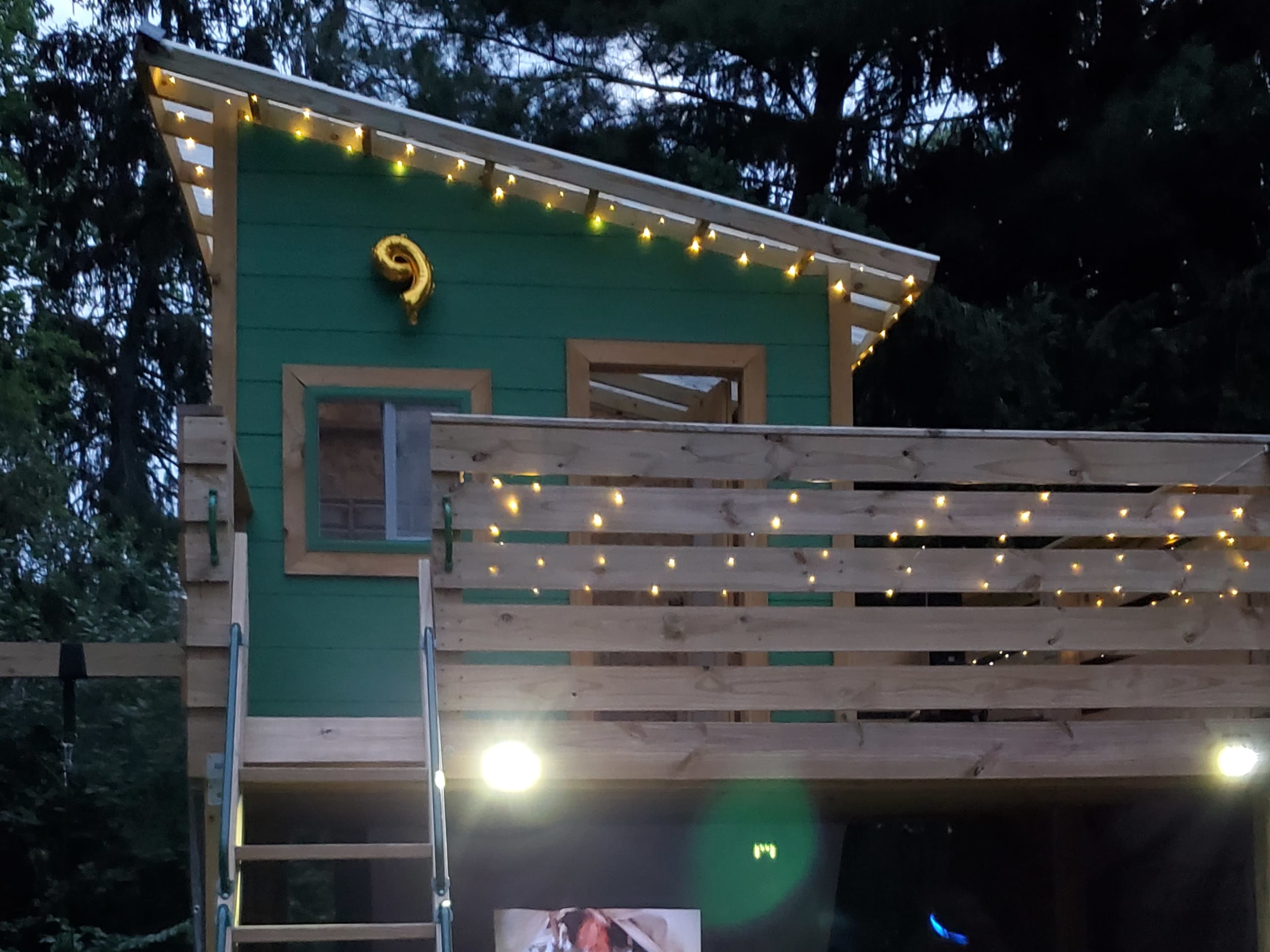 A small green treehouse with a wooden balcony, adorned with string lights, nestled among trees.