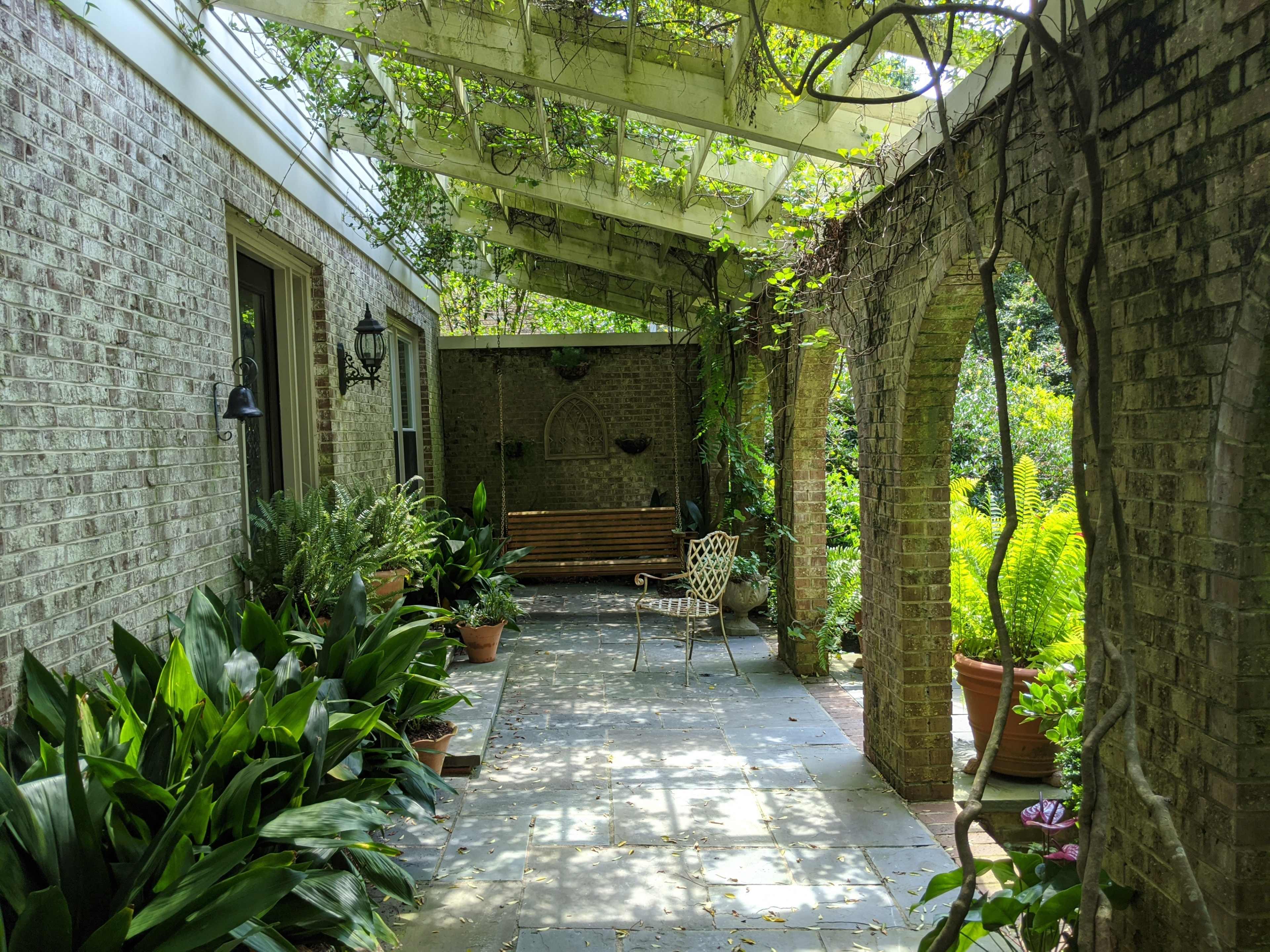 A stone pathway leads through an archway to a shaded outdoor seating area surrounded by potted plants and greenery.