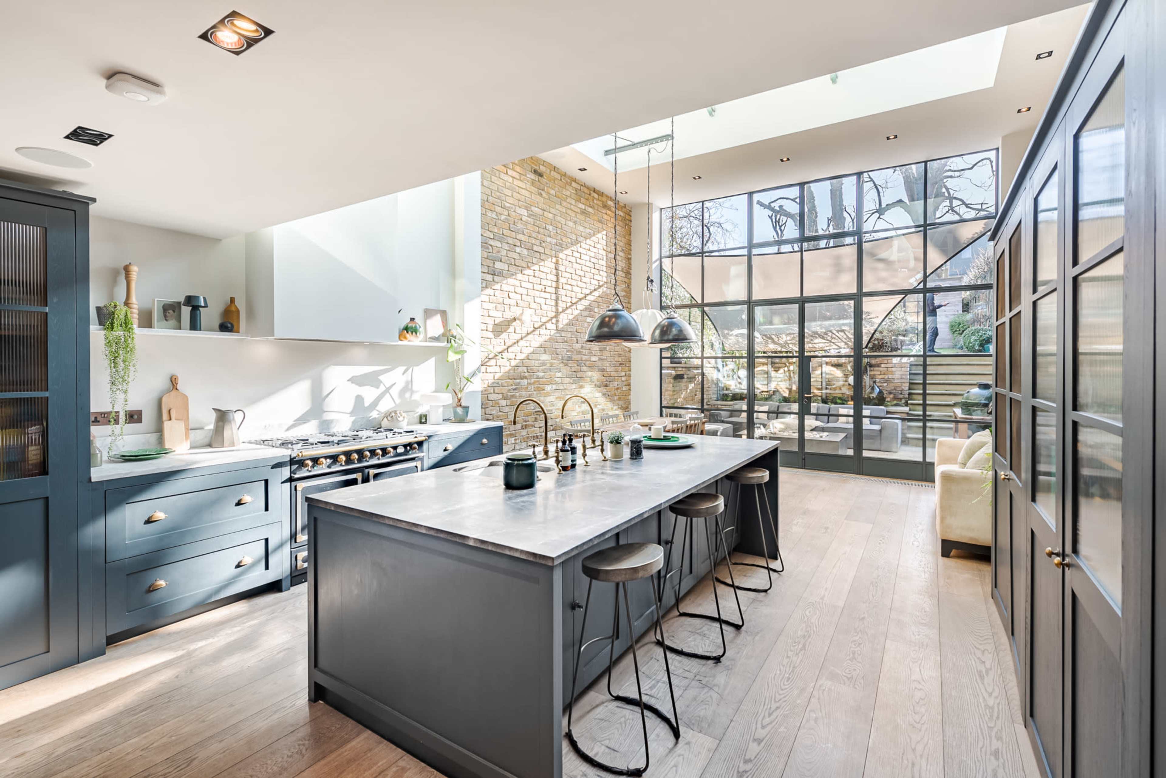 A modern kitchen with a large island, integrated appliances, and a glass wall that opens to a garden area.