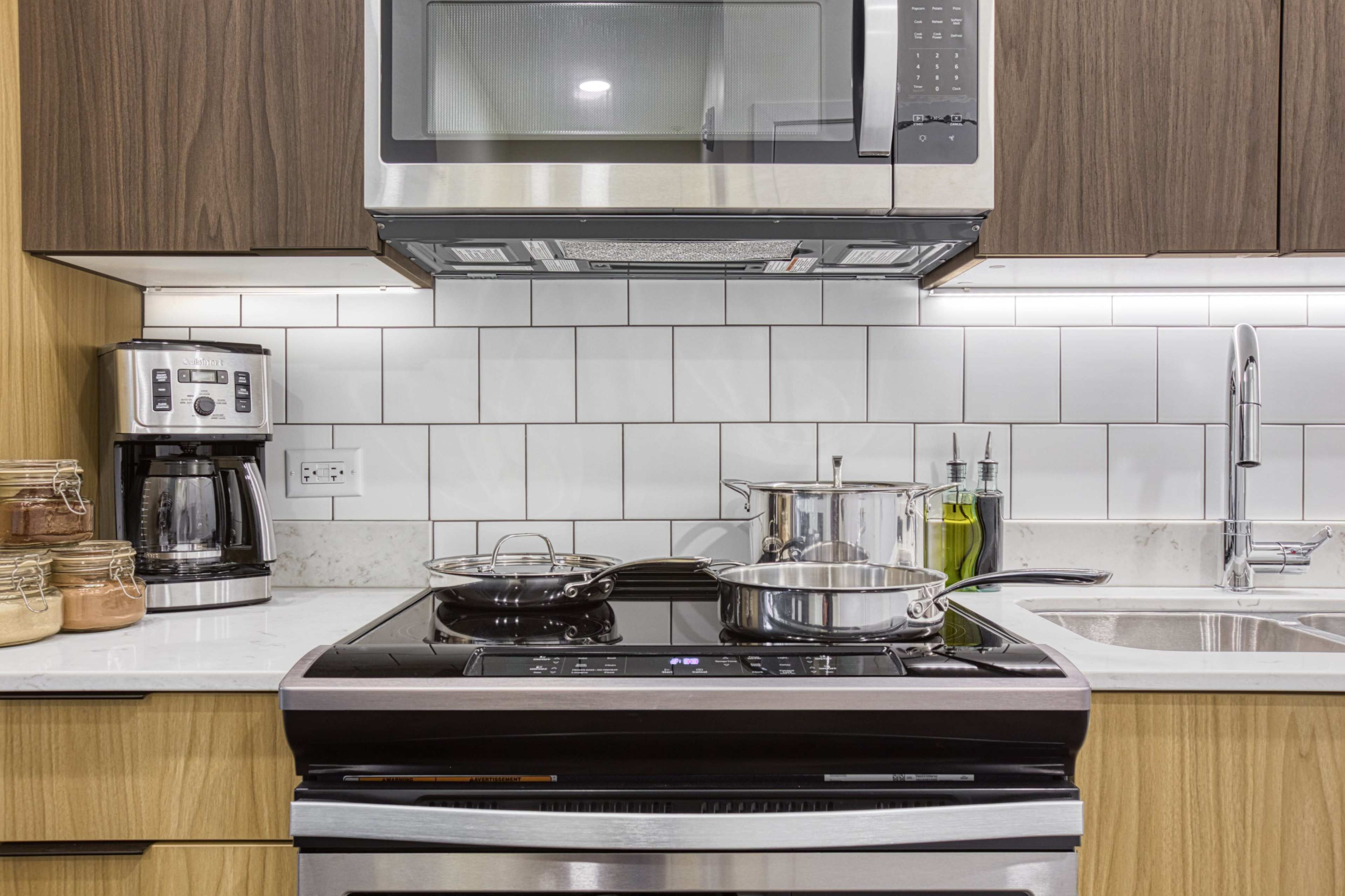 A modern kitchen countertop featuring a stove with pots, a microwave above, a coffee maker, and olive oil on the side.