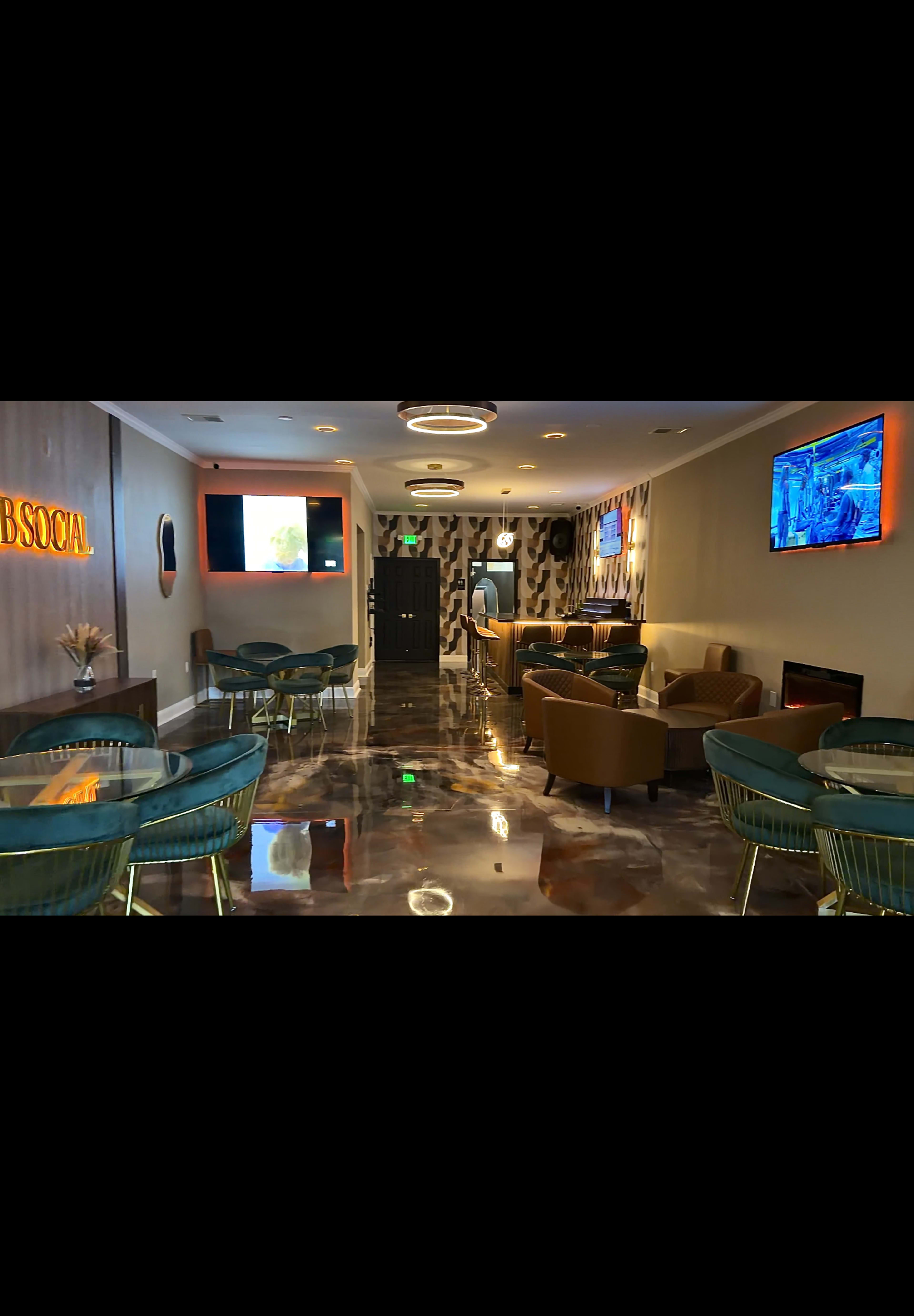 The image shows a modern lounge area featuring tables, chairs, and multiple screens on the walls.