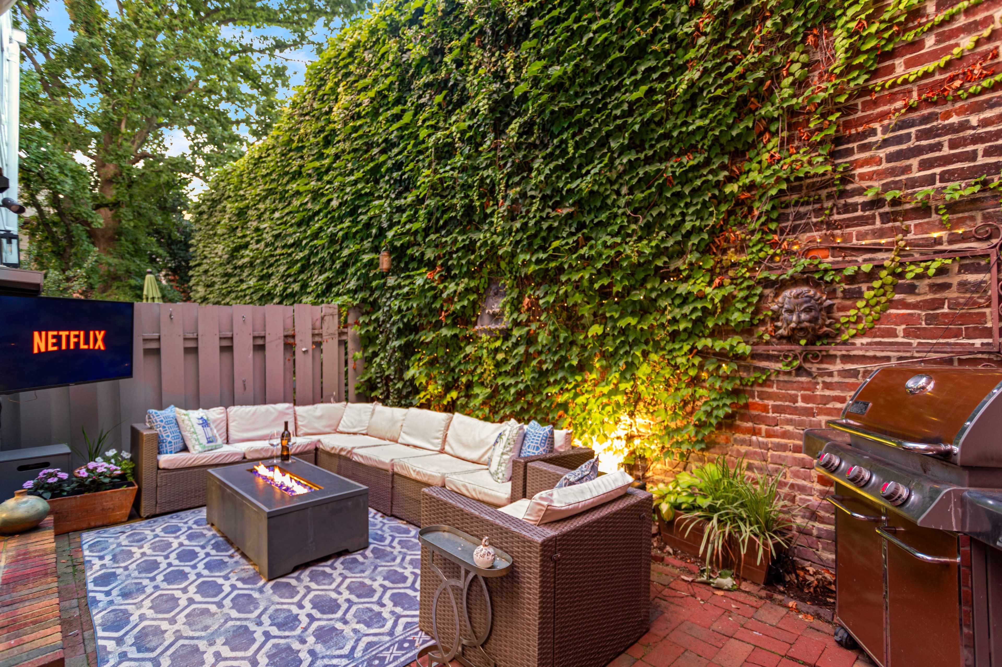 The image shows a cozy outdoor patio with a sectional sofa arranged around a fire pit, a grill on one side, and a wall covered in greenery.