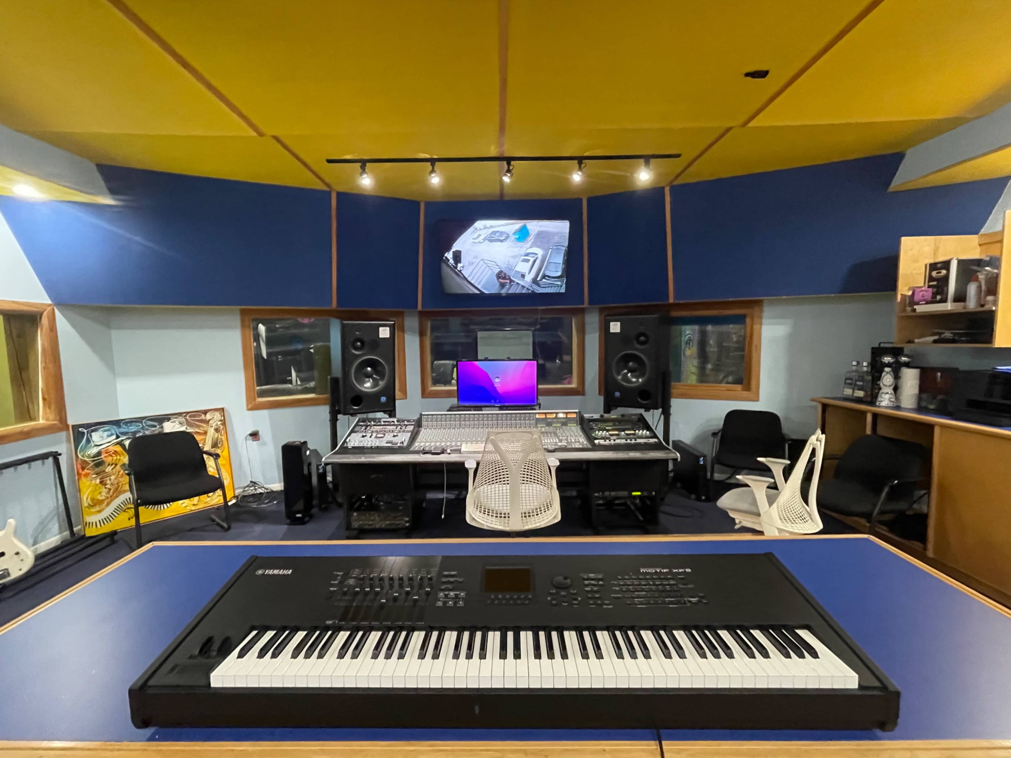 The image shows a recording studio with a digital keyboard in the foreground, a mixing console, speakers, and a computer monitor in the background.