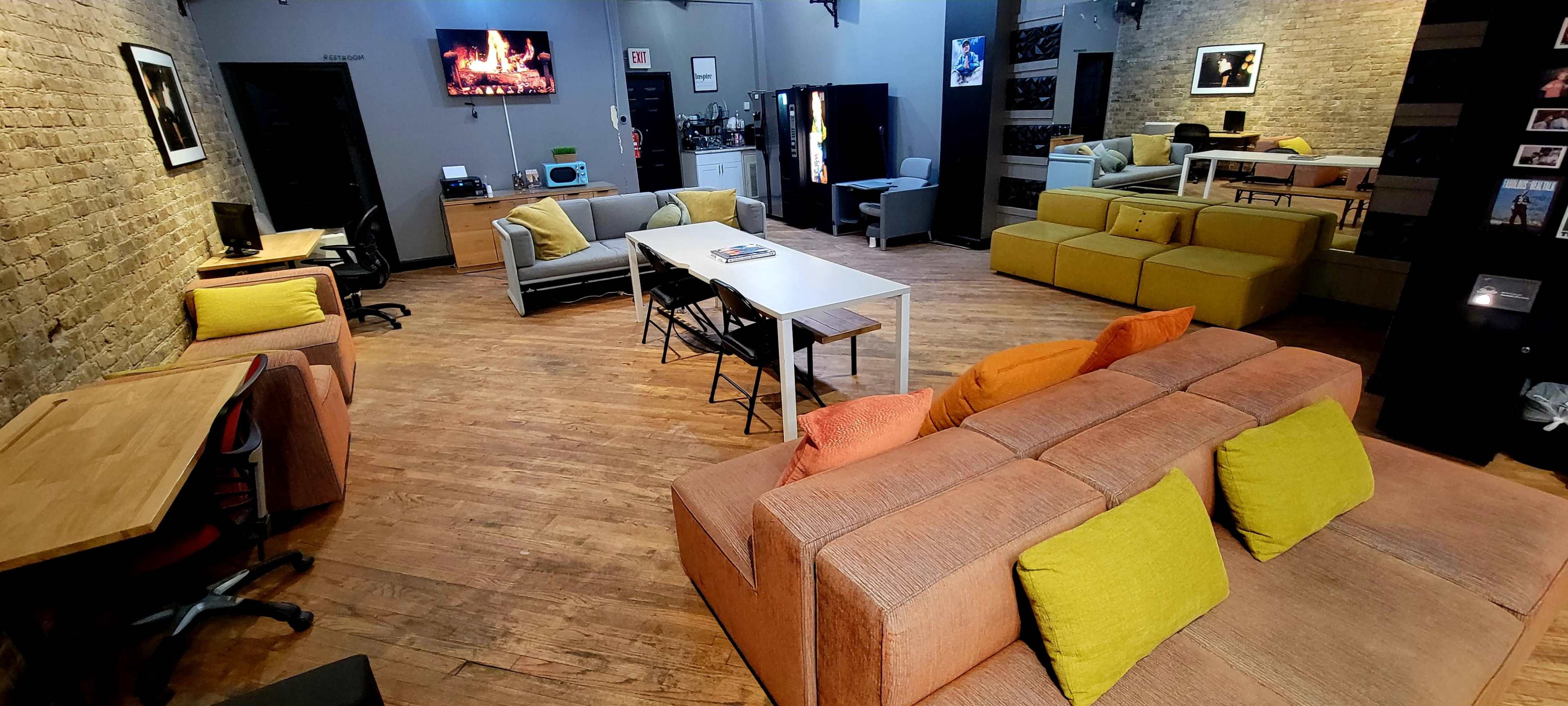 The image shows a spacious common area with wooden flooring, various seating arrangements including sofas and tables, and a kitchenette in the background.