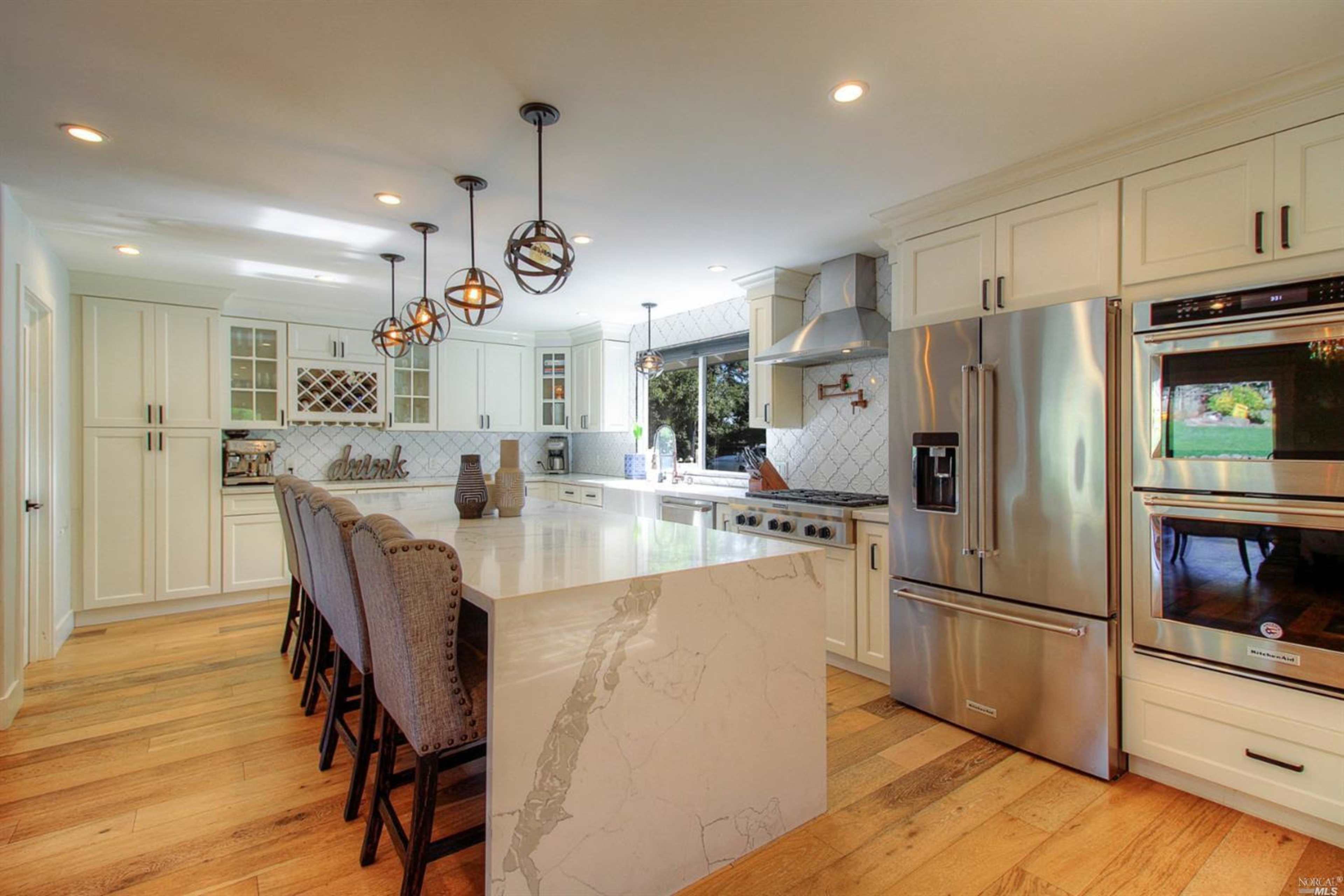 A modern kitchen features white cabinetry, a large marble island with seating, and stainless steel appliances.