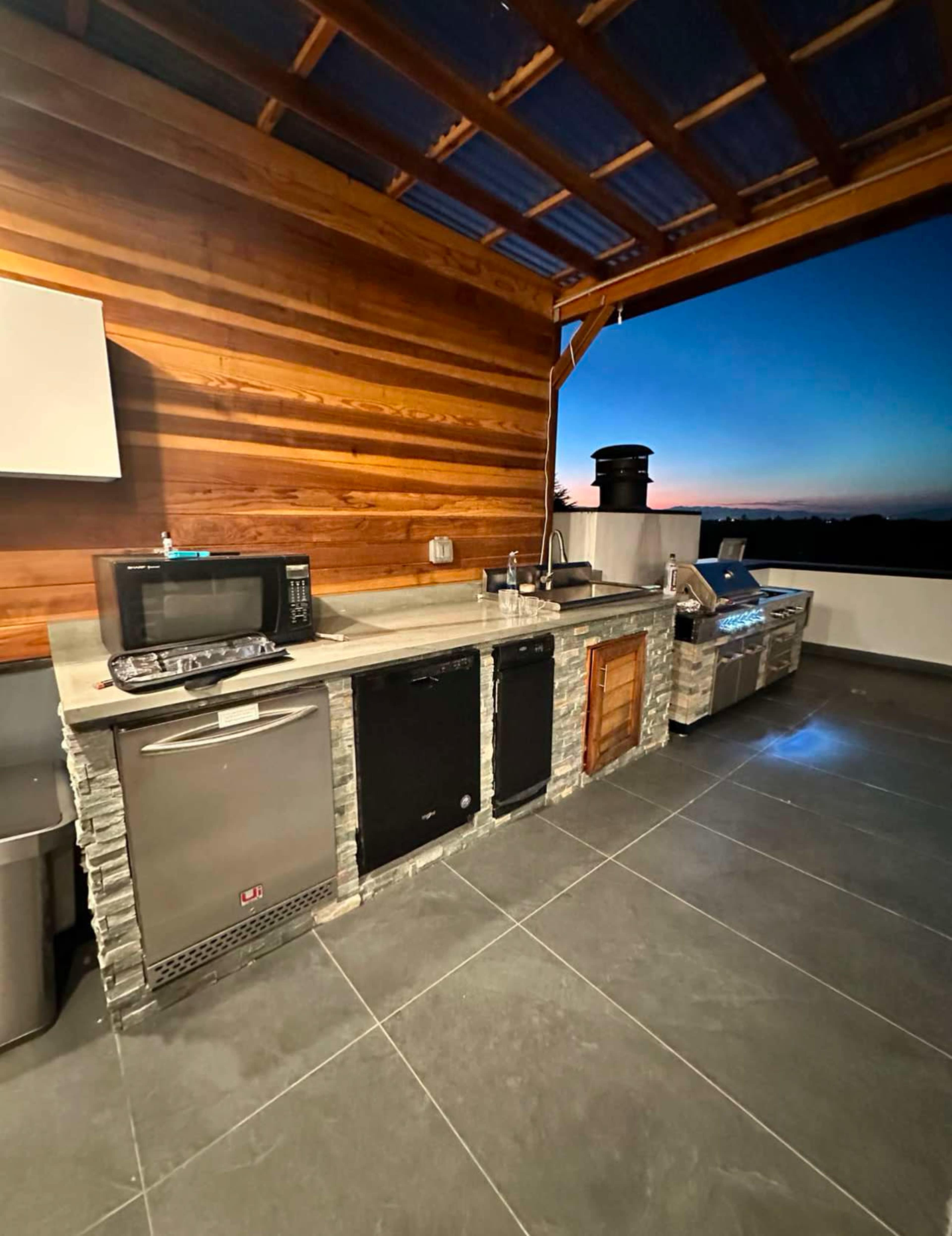 The image shows an outdoor kitchen with a stone countertop and several appliances including a microwave, refrigerator, and grill, under a wooden overhang at sunset.
