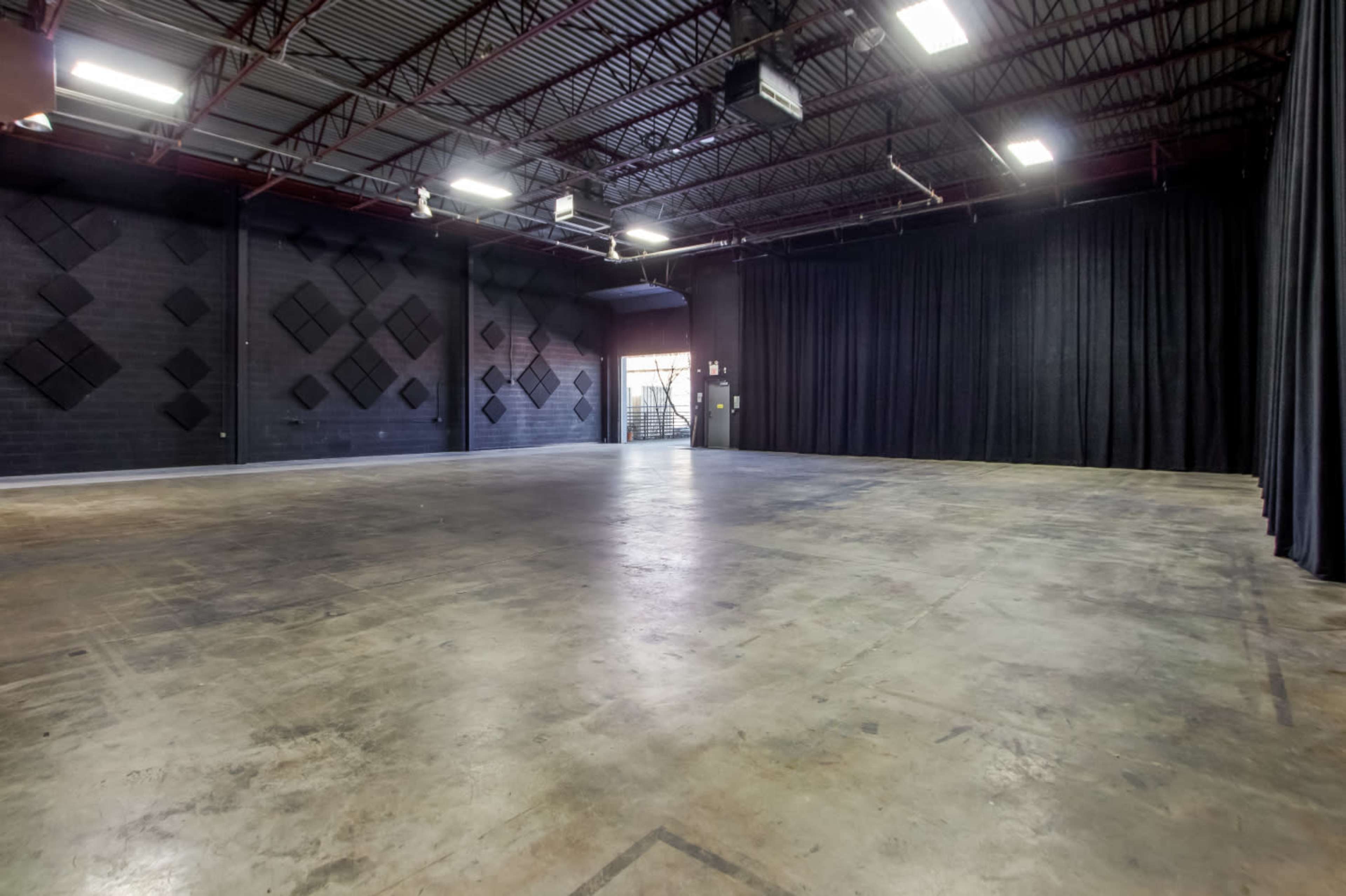 A large, empty room with a polished concrete floor, black walls featuring sound panels, and a set of double doors at the back.