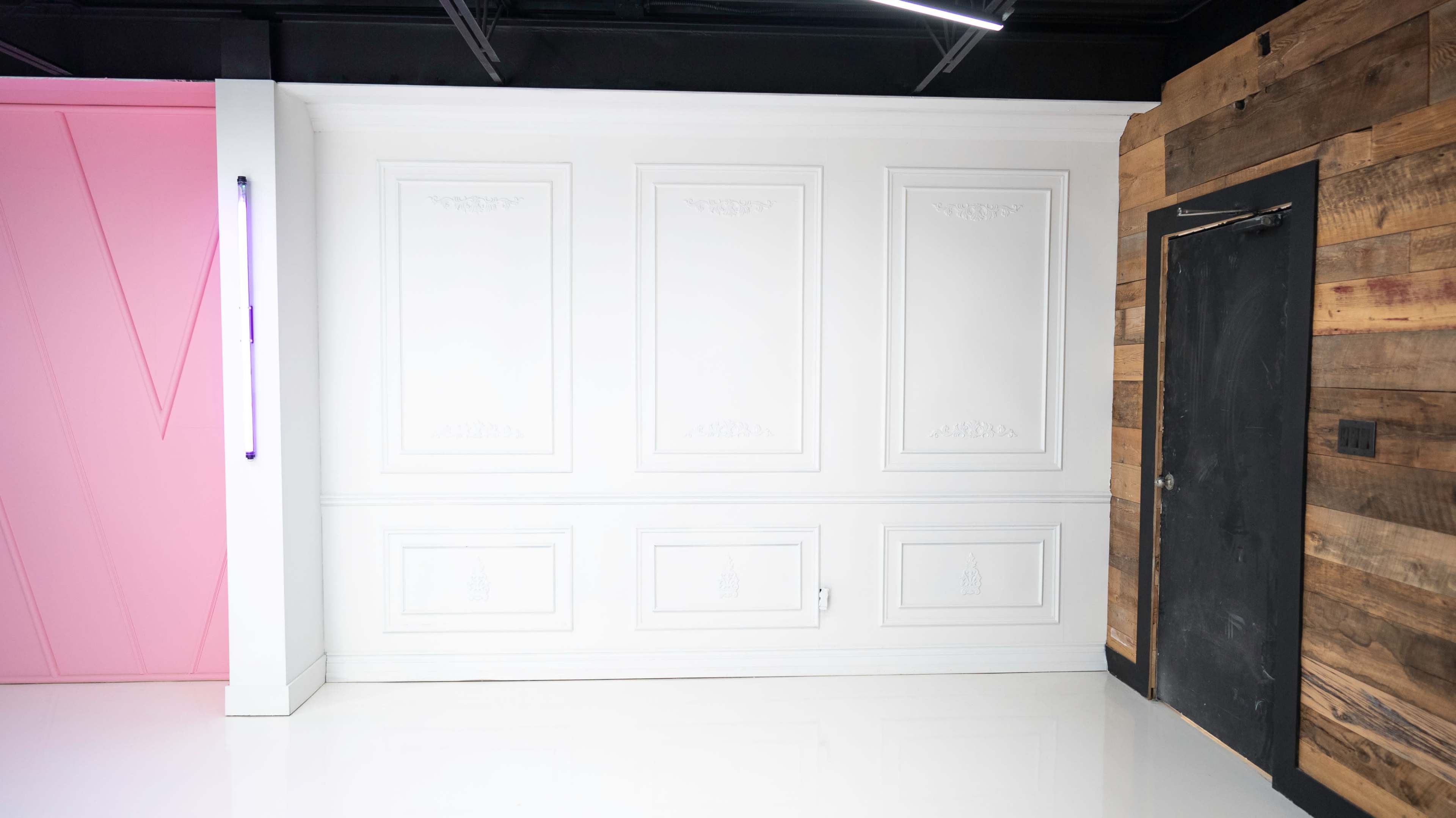A smooth, white wall with decorative paneling next to a rustic wooden wall and a black door.